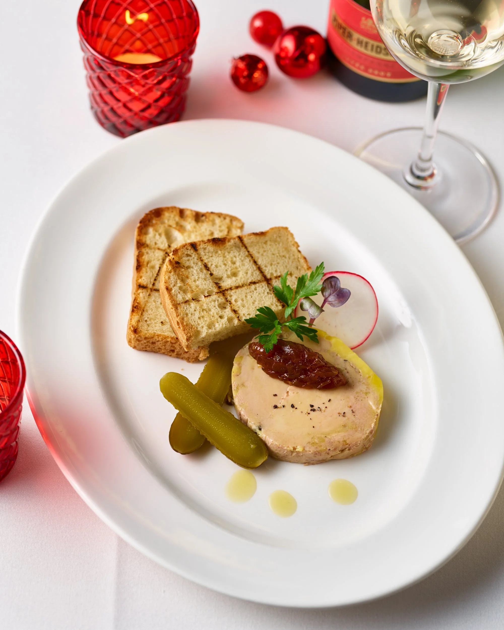 A white plate with slices of toasted bread, green pickles, and a pâté topped with a dollop of chutney, garnished with radish and herbs, with a glass of white wine and a red candle in the background.