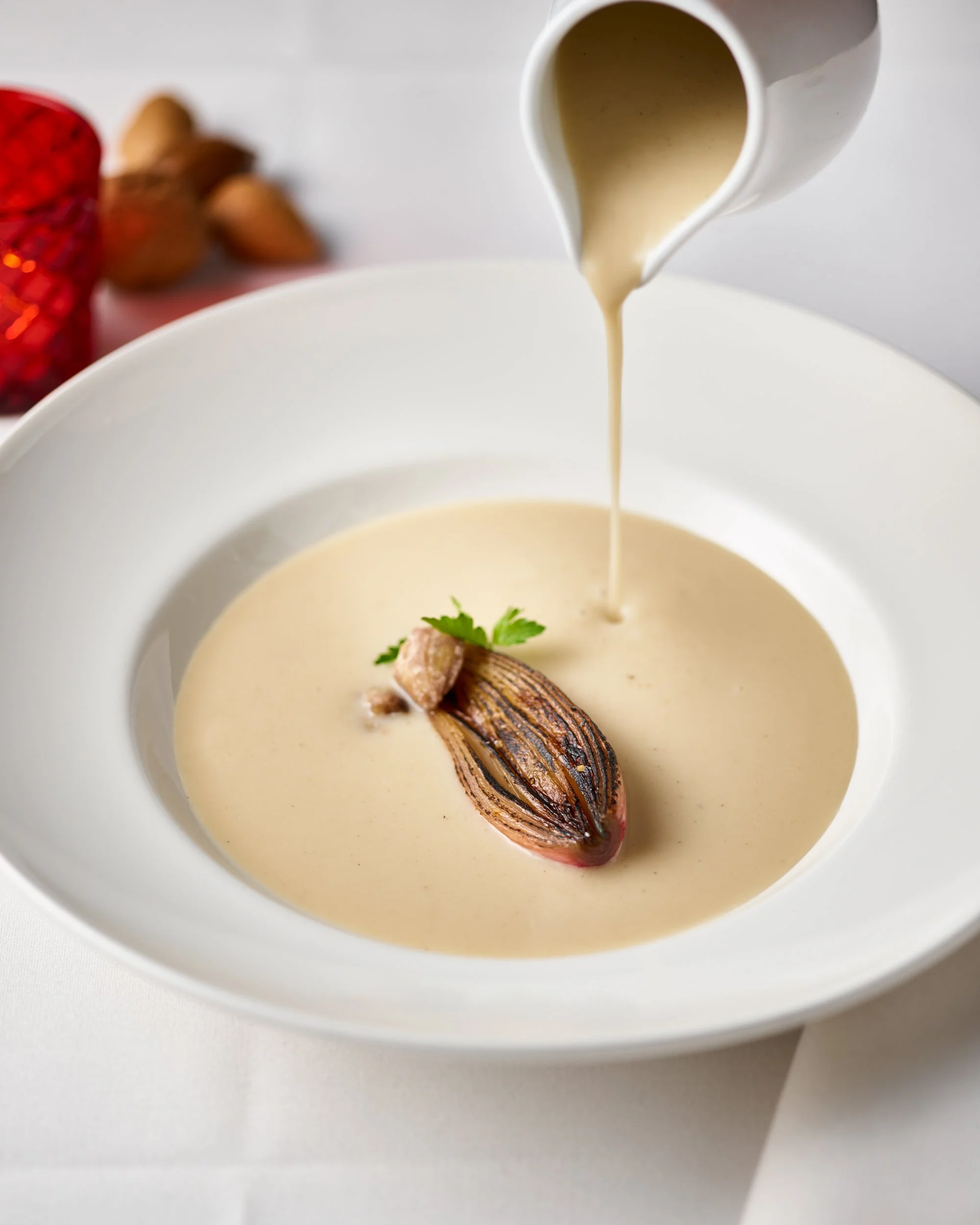 Creamy soup being poured into a white bowl, garnished with a grilled onion and a small green herb, with a background of nuts and red decorative items.