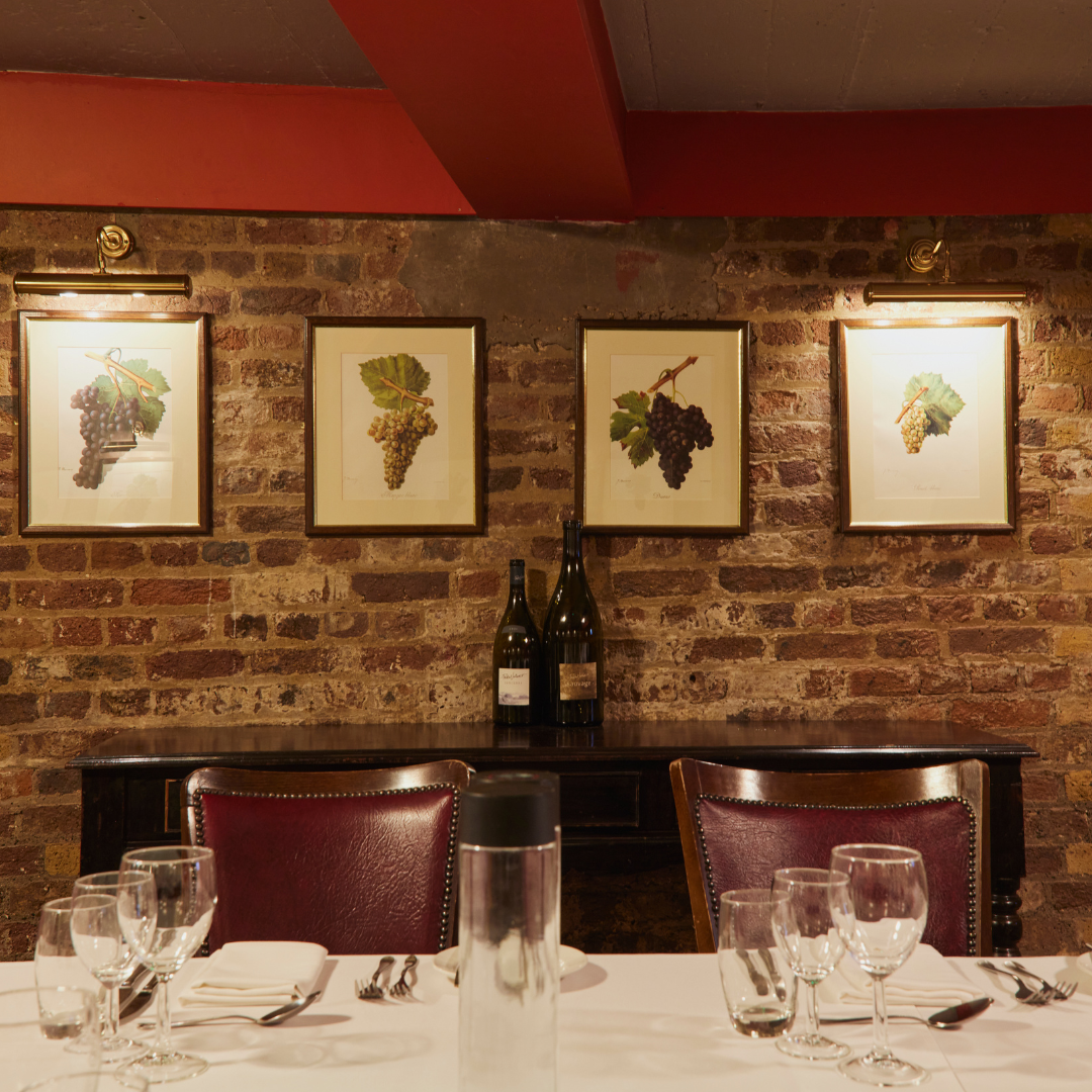 A restaurant dining area with a rustic brick wall decorated with framed grapevine illustrations, two wine bottles on a dark wooden sideboard, and a table set with glasses, silverware, napkins, and a water pitcher.