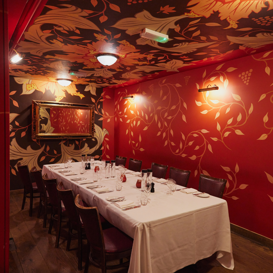 Elegant dining room with a long, white tablecloth-covered table set for a meal, surrounded by dark wooden chairs. The room has vibrant red walls with leaf patterns and a mirror on one wall. The ceiling features floral and leaf designs, with lighting fixtures providing warm illumination.