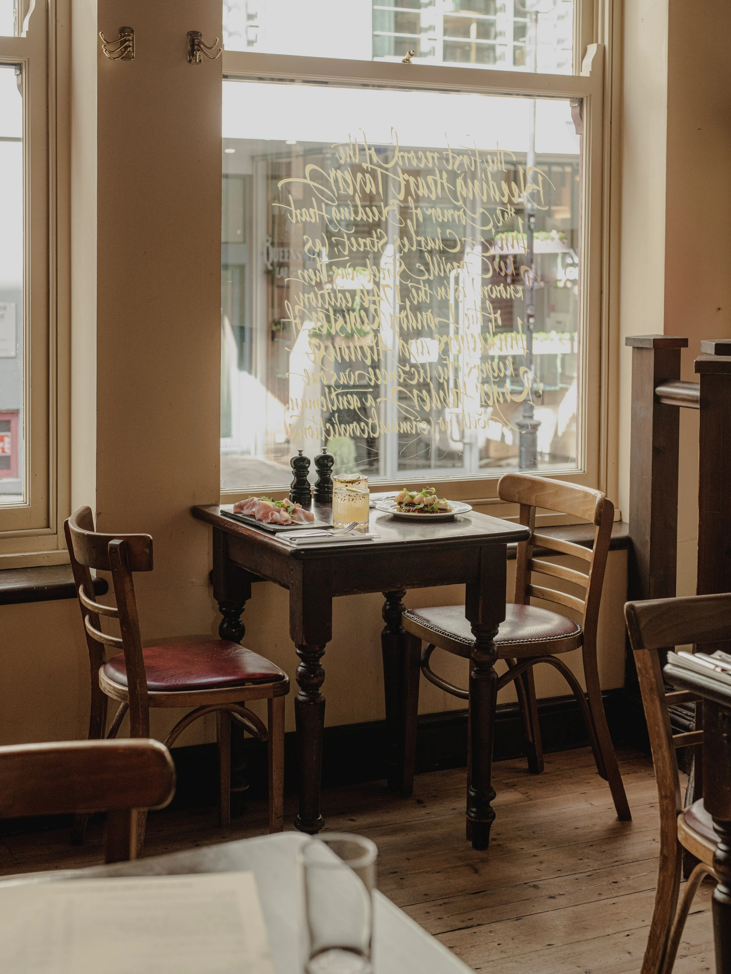 A cozy restaurant corner with a wooden table set with food and drinks, surrounded by wooden chairs, near a large window with handwritten decorative gold script on it, letting in natural light.