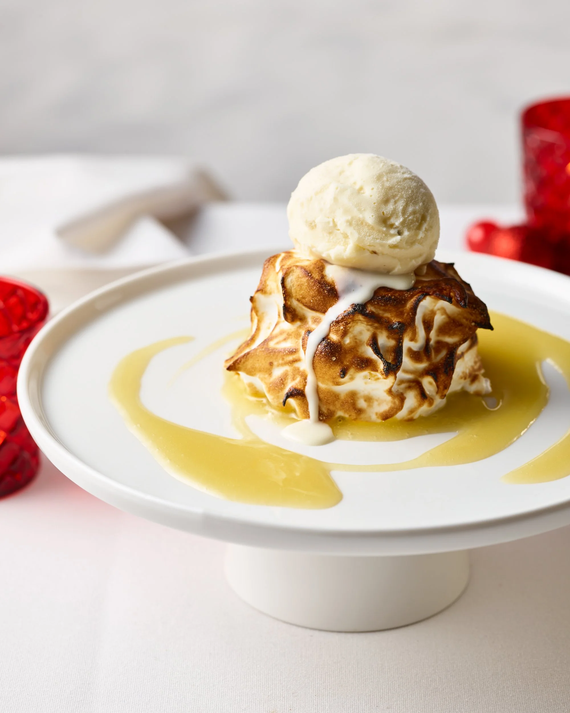 A dessert with a toasted meringue topping, a scoop of vanilla ice cream, and drizzled with honey on a white cake stand.