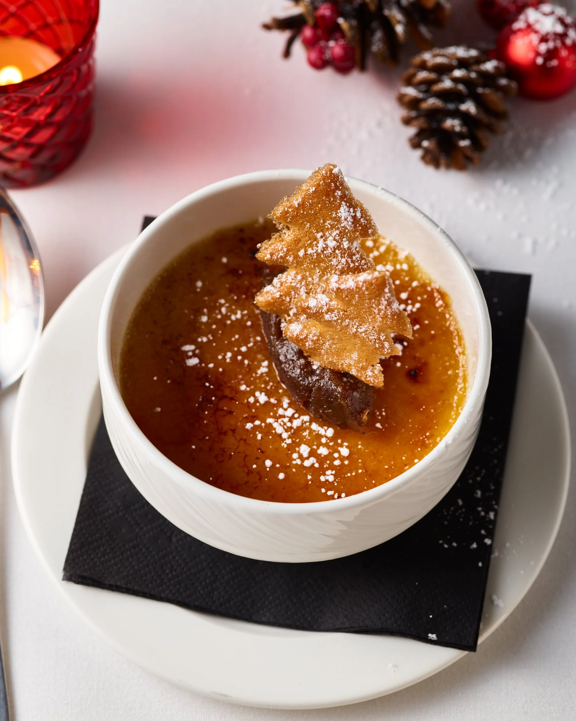 Creamy caramel crème brûlée topped with a decorative caramelized sugar tree, served in a white ramekin on a black napkin with holiday decorations in the background.