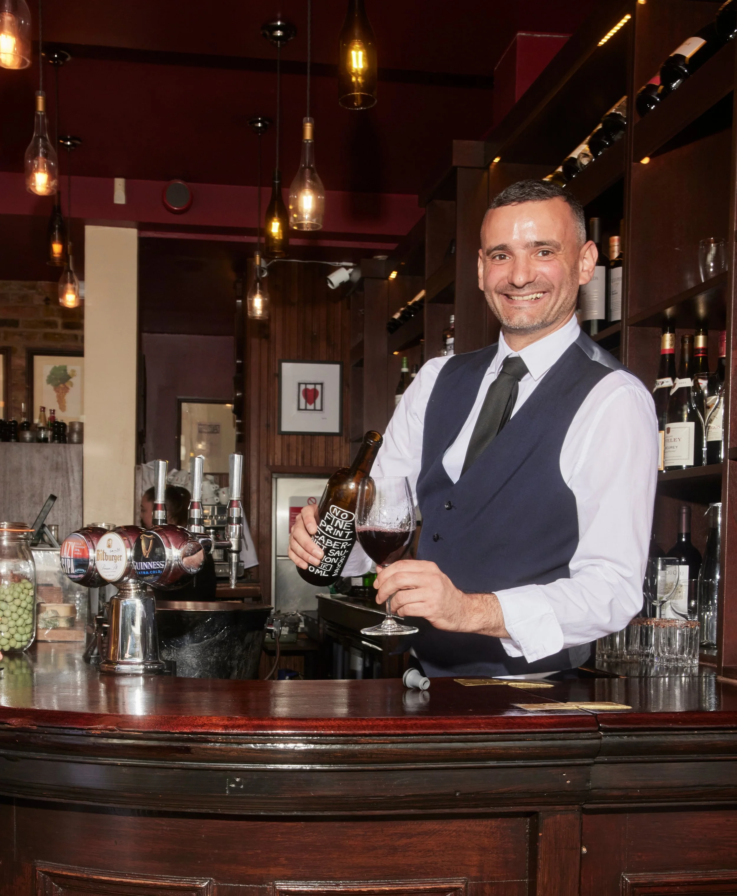 A smiling bartender in a vest pours red wine at a bar with bottles and taps behind him.