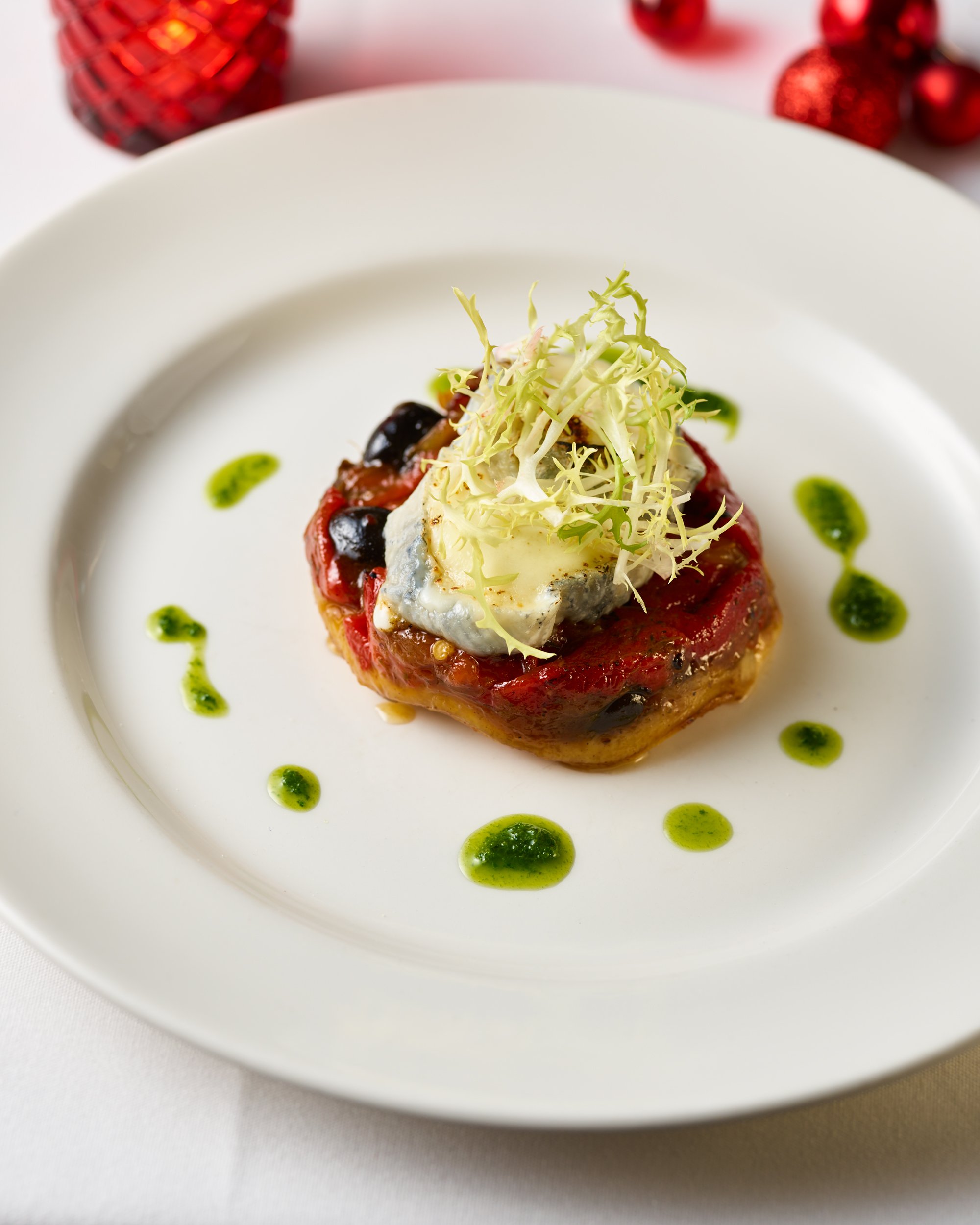 A gourmet appetizer on a white plate featuring a baked tomato with a topping of cheese, garnished with shredded lettuce and drizzled with green sauce.