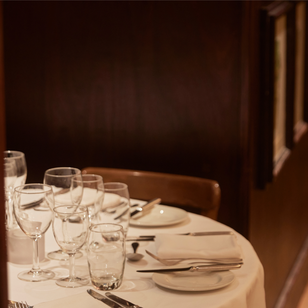 A round dining table set with multiple wine glasses, water glasses, plates, napkins, knives, and forks in a cozy restaurant or dining room.