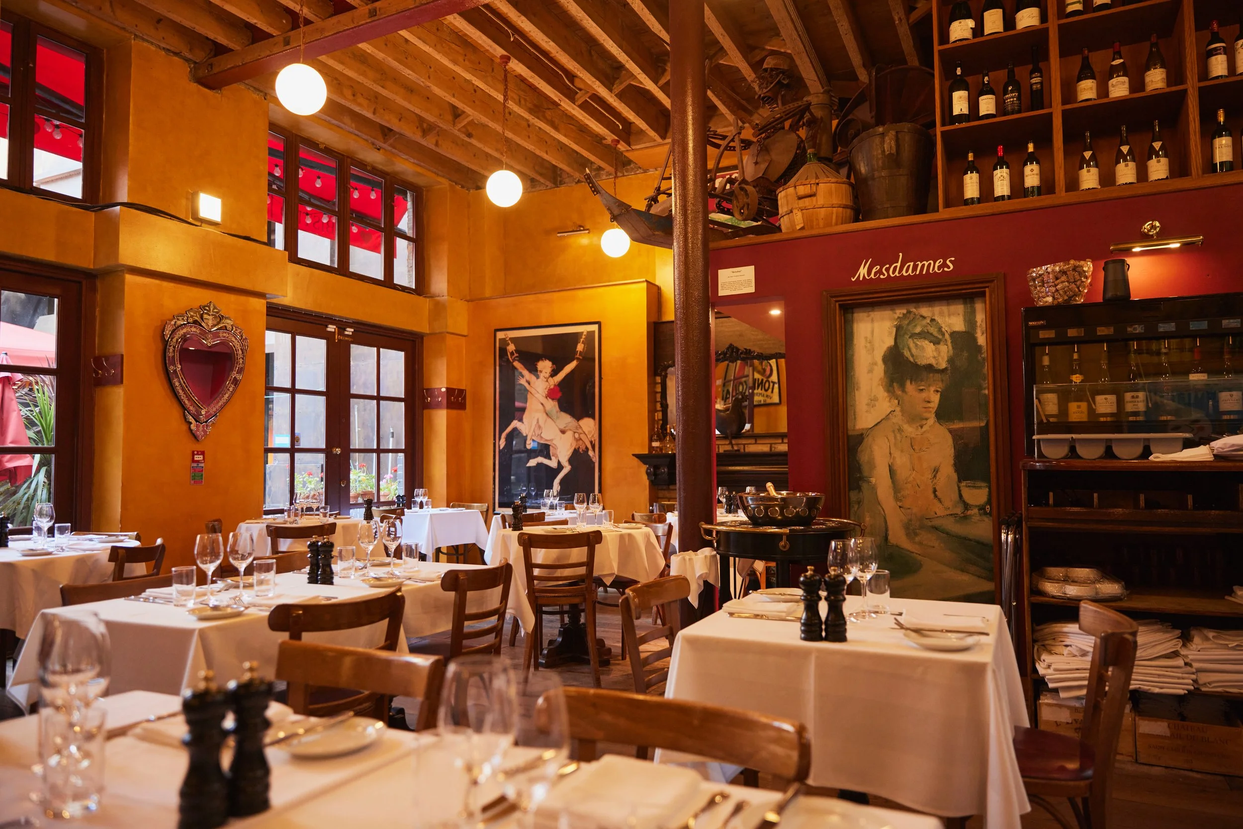 Interior of a cozy Italian restaurant with yellow and red walls, wooden ceiling beams, and large windows with red trim. The dining area has tables covered with white tablecloths, set with wine glasses, plates, and cutlery. Artwork and vintage decor, 