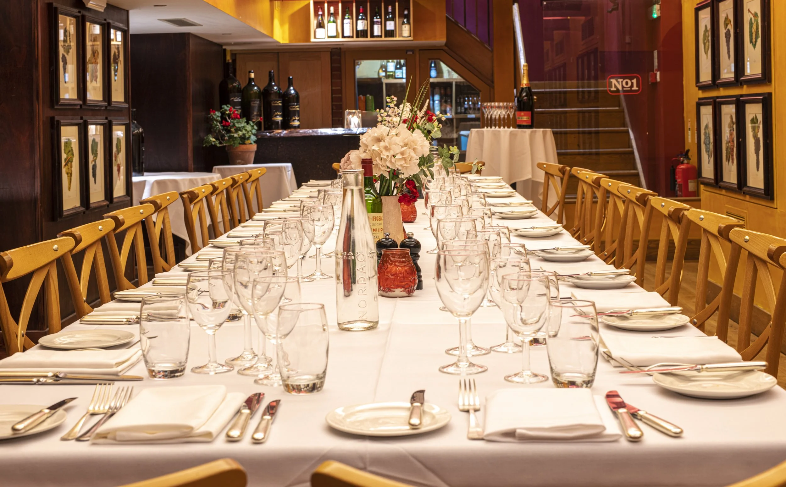 A long dining table set for a meal in a restaurant or banquet hall, with white tablecloth, neatly arranged plates, silverware, glasses, and floral centerpieces, surrounded by wooden chairs.