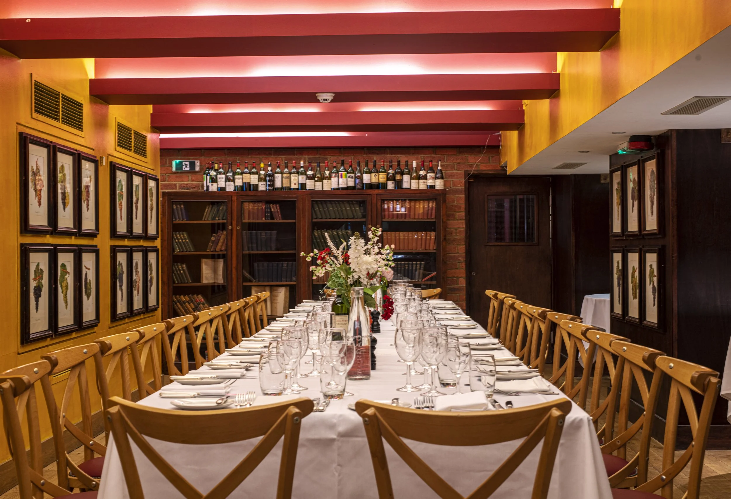 A long dining table set for a formal dinner with white tablecloths, glassware, and napkins, in a restaurant with a brick wall, wooden shelves with wine bottles, framed artwork of grapes, and a floral centerpiece.