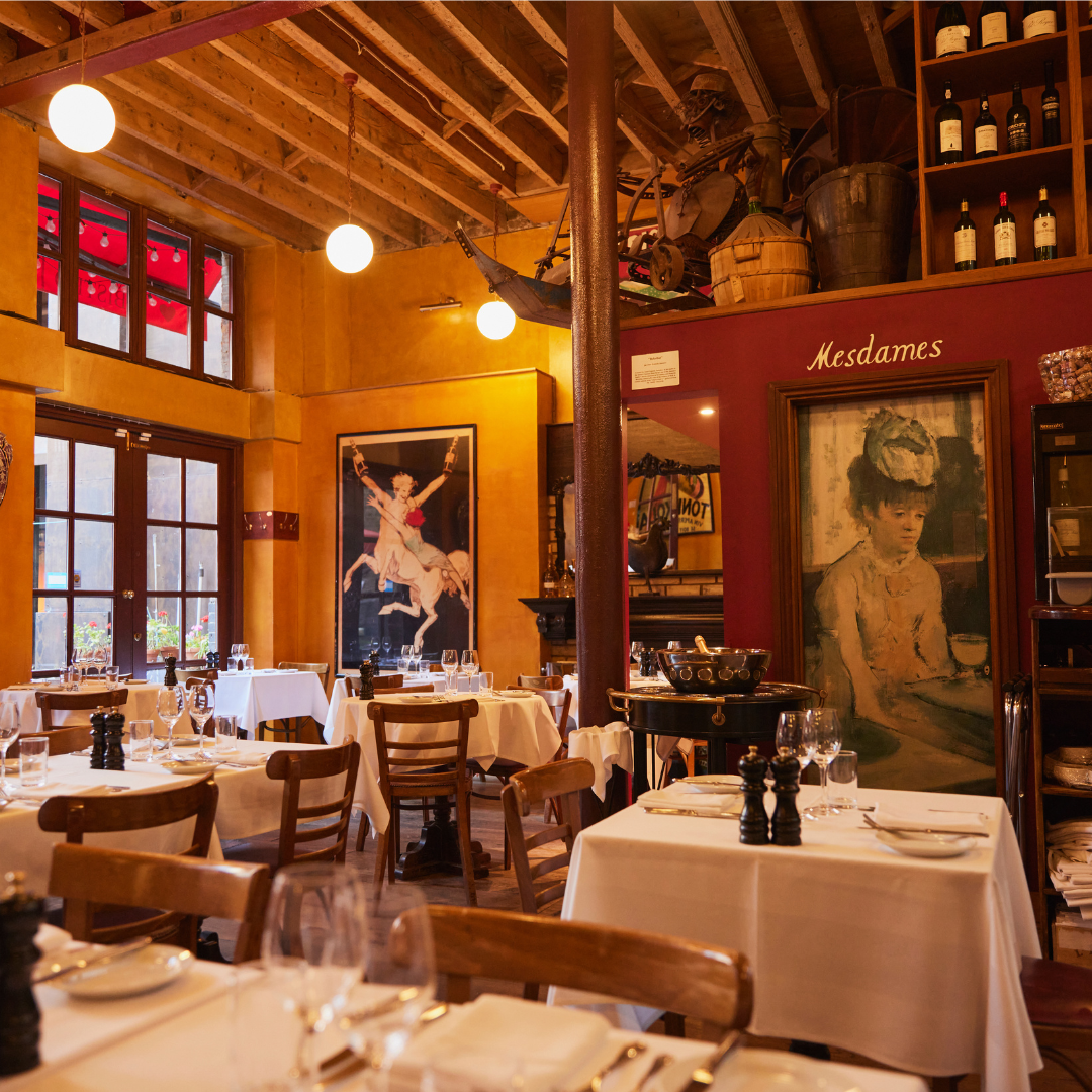 Interior of a warmly lit restaurant with wooden chairs and white tablecloths, decorated with vintage artwork and bottles on shelves.