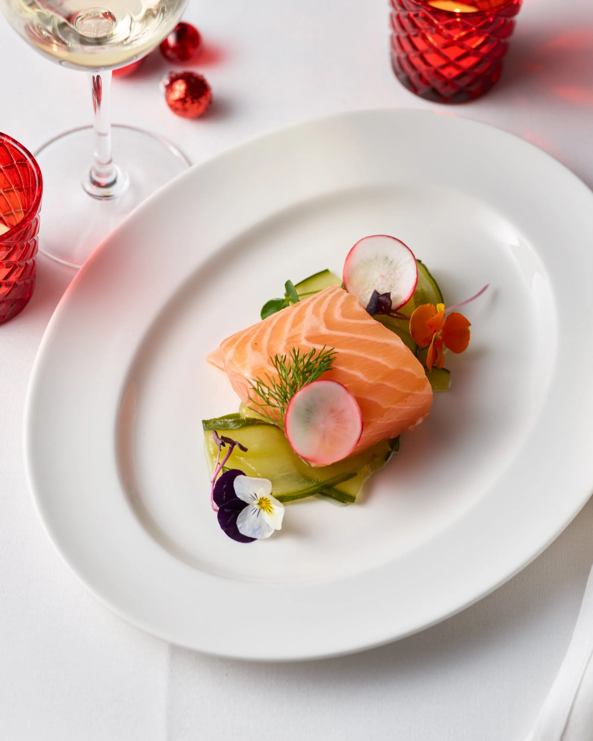 Plate with a piece of cooked salmon garnished with radish slices, cucumber, herbs, and edible flowers, set on a white table with a wine glass and decorative glassware in the background.
