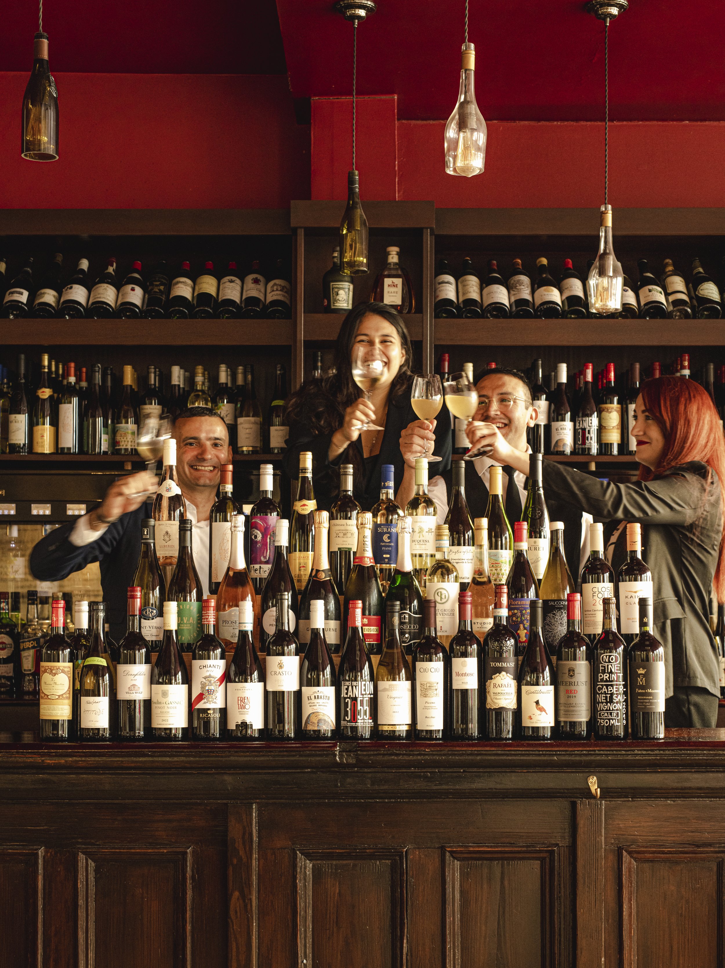 People celebrating and raising glasses of wine at a bar with a large selection of wine bottles.