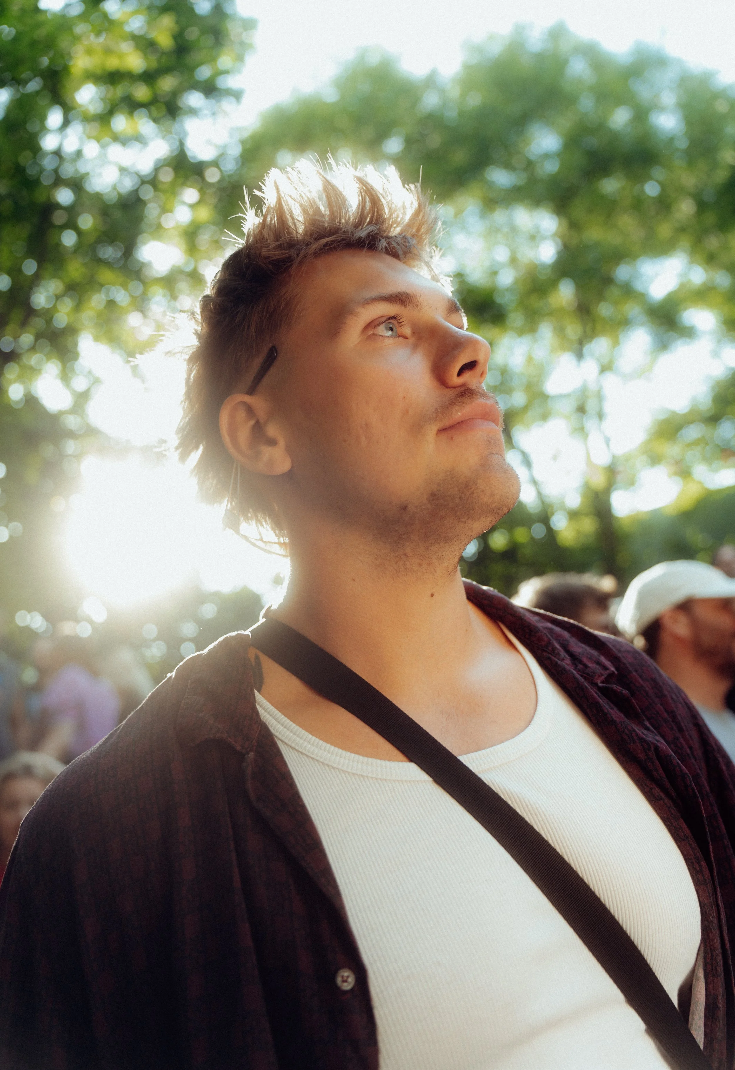 Junger Mann mit kurzen, gefärbten Haaren, schaut nach oben, Sonnenlicht hinter ihm, im Park mit grünen Bäumen, trägt weiße T-Shirt und schwarze Schultertasche.
