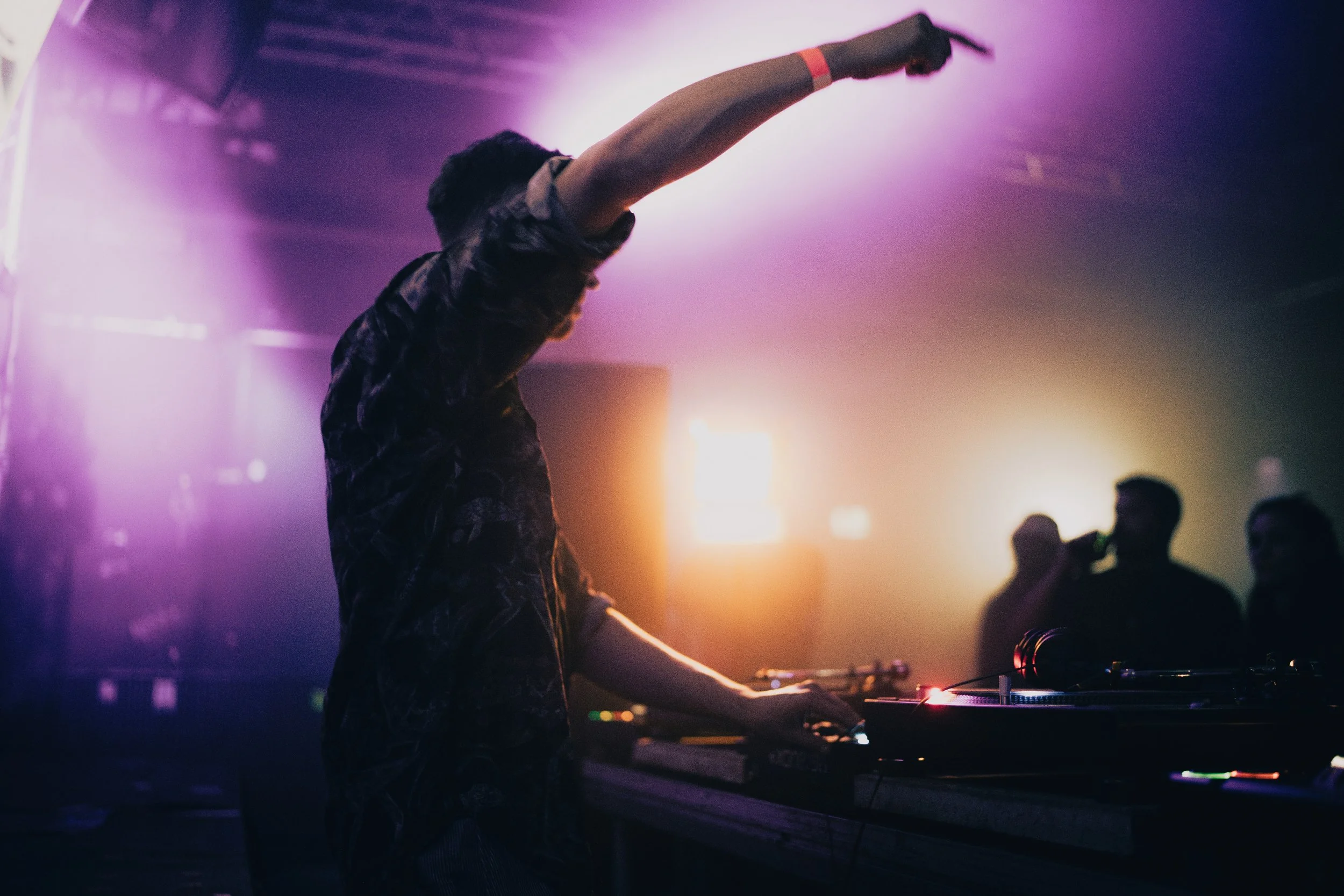 Ein DJ spielt an einem DJ-Pult in einem dunklen Club mit buntem Licht.