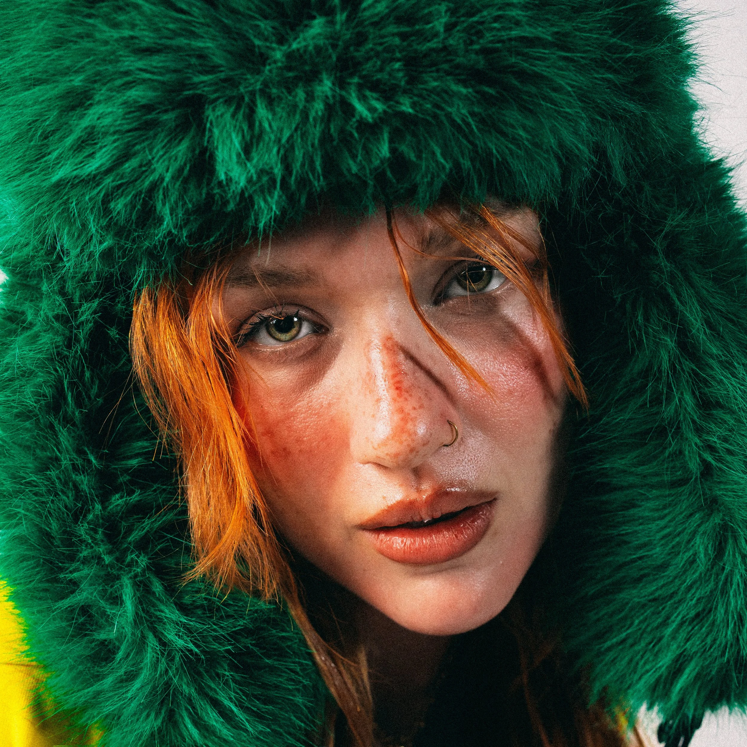 Nahaufnahme eines Gesichts mit grüner Fellkapuze, Rothaarige mit Sommersprossen, Nasenpiercing, roter Haarspitze, natürlichem Make-up und leicht geöffneten Lippen.