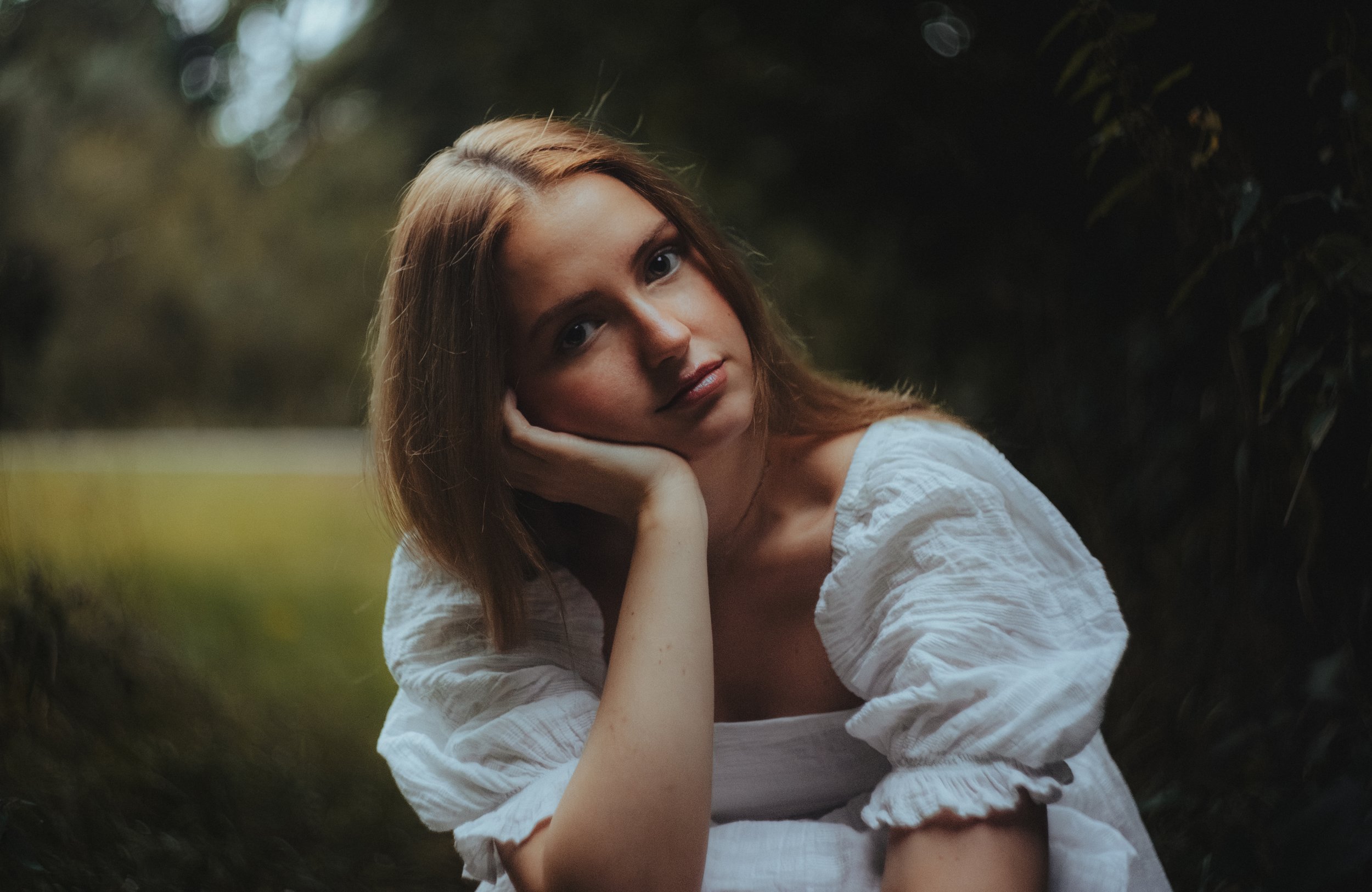 Junge Frau mit braunen Haaren in einem weißen Kleid sitzt im Grünen, stützt ihren Kopf mit der Hand und schaut nachdenklich in die Kamera.