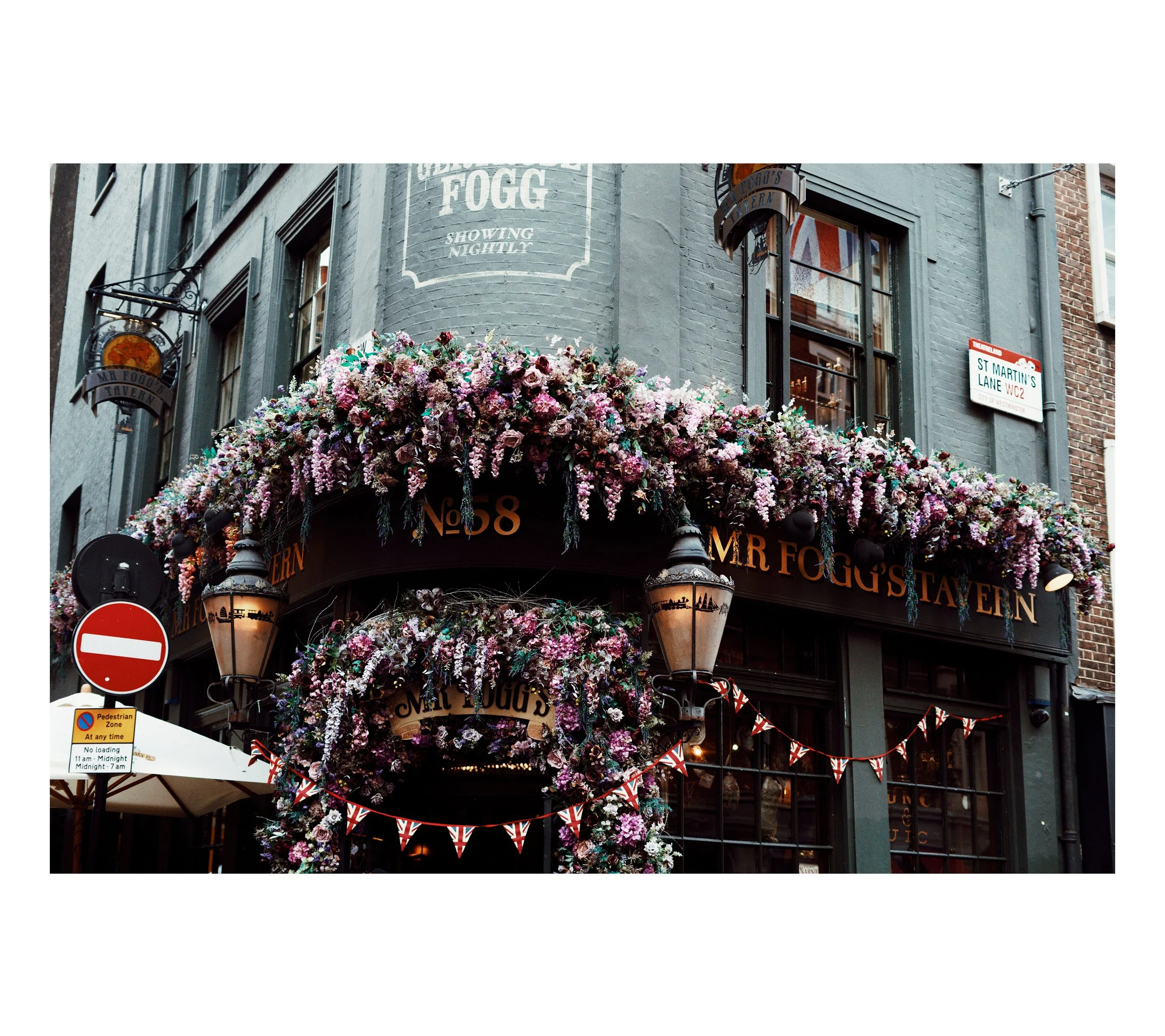 Außenansicht eines Pubs mit vielen Blumen und Flaggen, Pag-schild inklusive.