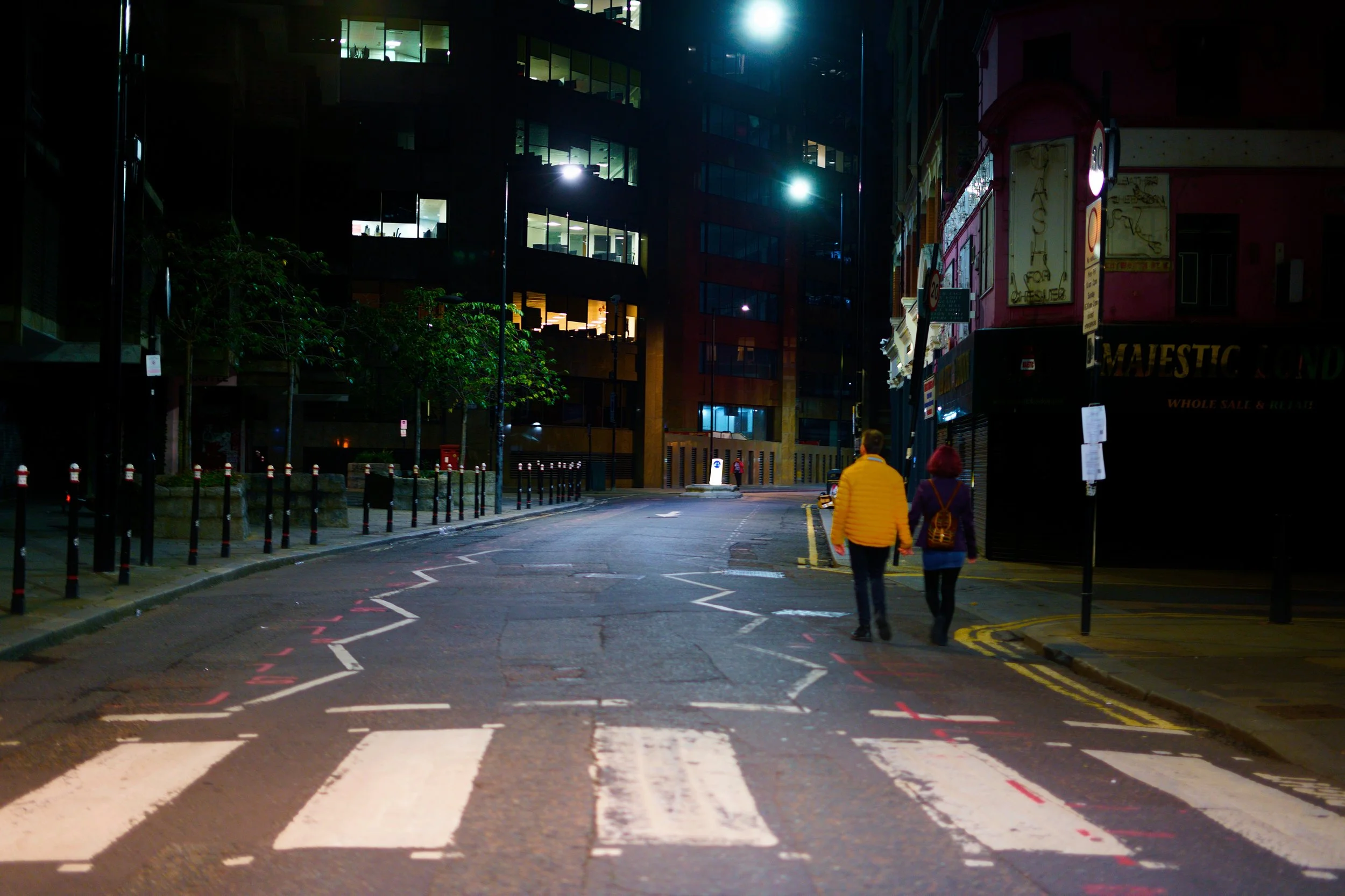 Zwei Personen gehen nachts in einer Stadtstraße, umgeben von modernen Gebäuden und Straßenlaternen.