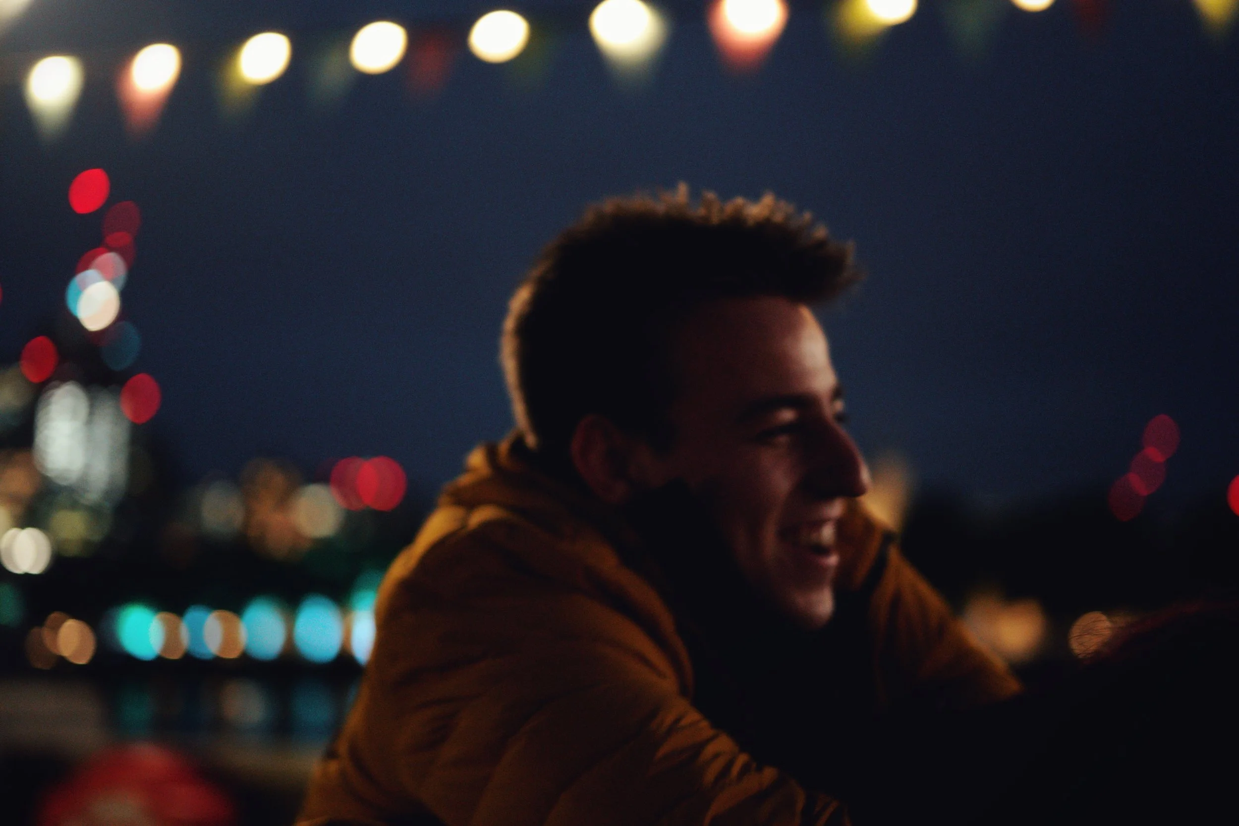 Ein lachender junger Mann in einem braunen Jacke, der bei Nacht draußen ist, mit bunten Lichter im Hintergrund.
