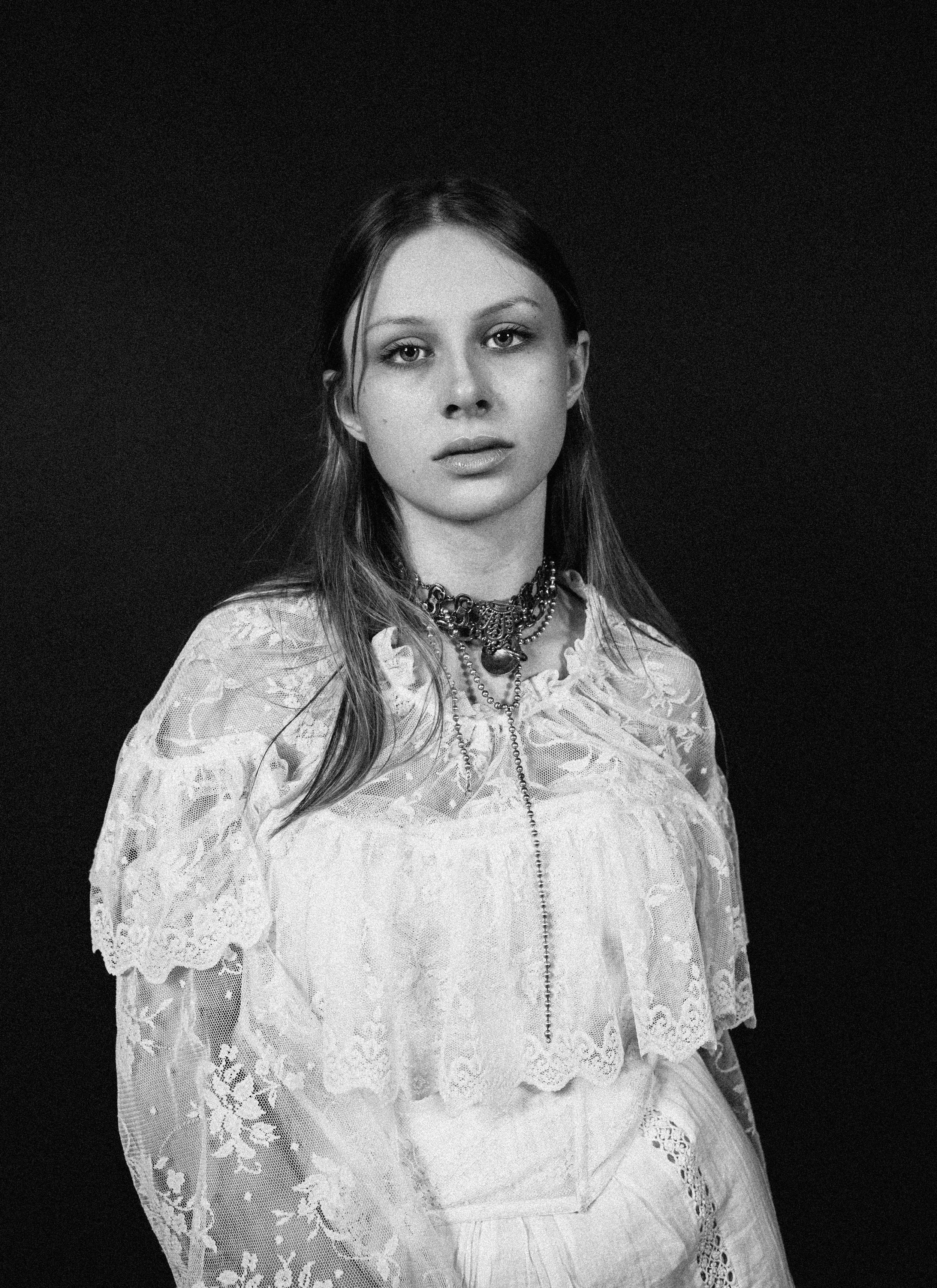 Schwarz-weiß-Fotografie eines jungen Mädchens mit langen Haaren, das direkt in die Kamera schaut, getragen in einem historischen, Spitzenkleid und mehreren Halsketten gegen einen schwarzen Hintergrund.