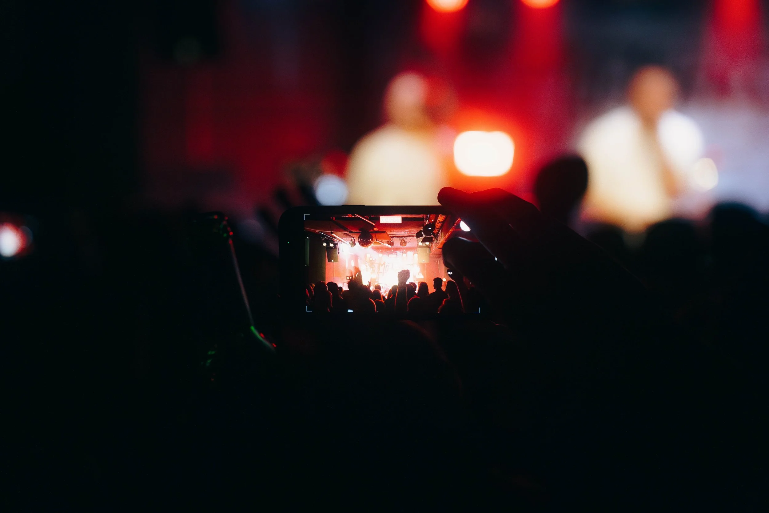 Menschen machen Fotos von einer Bühne bei einem Konzert, im Hintergrund sind Scheinwerfer und zwei Personen auf der Bühne zu sehen.