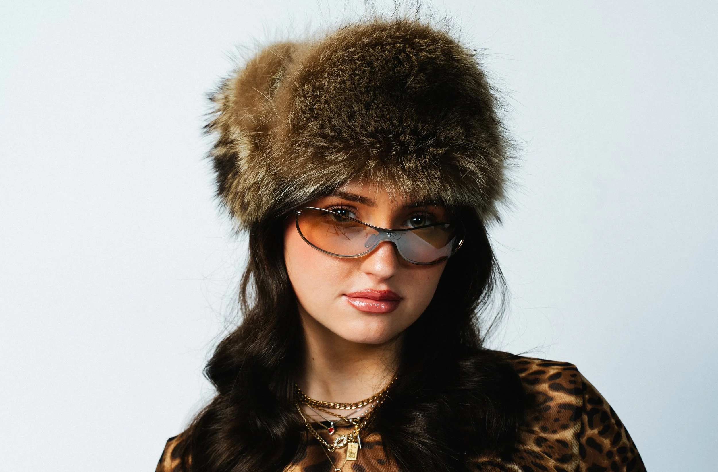 Junge Frau trägt einen Pelzmütze, Sonnenbrille, Tierprint-Bluse und mehrere Halsketten.