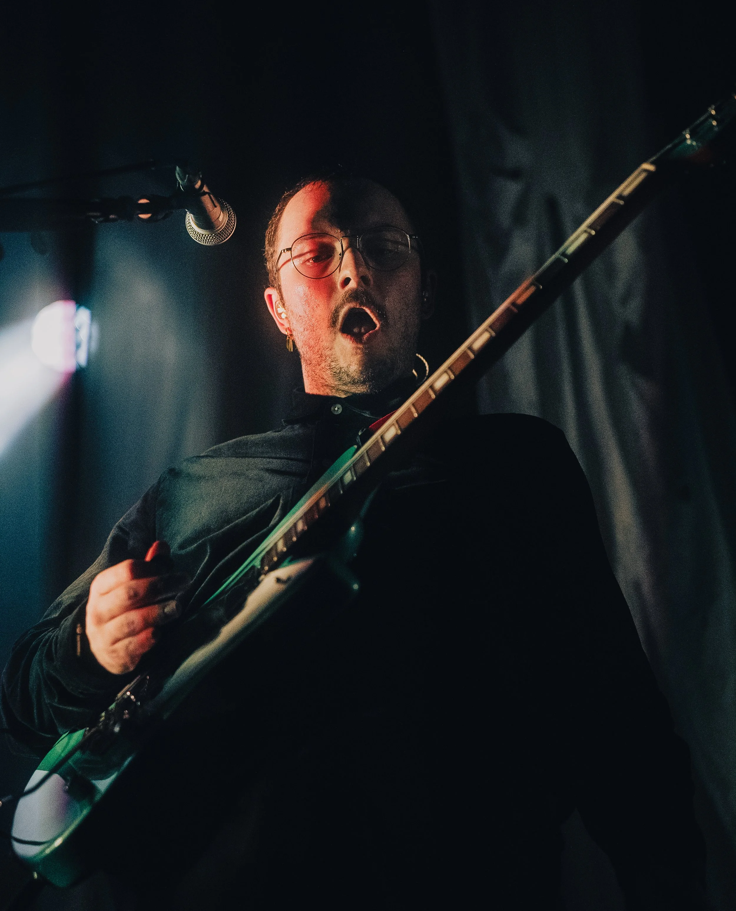 Ein Mann mit dunklem Hemd spielt E-Gitarre bei einem Auftritt, er trägt Brille und hat kurze Haare, im Hintergrund dunkler Bühnenhintergrund.