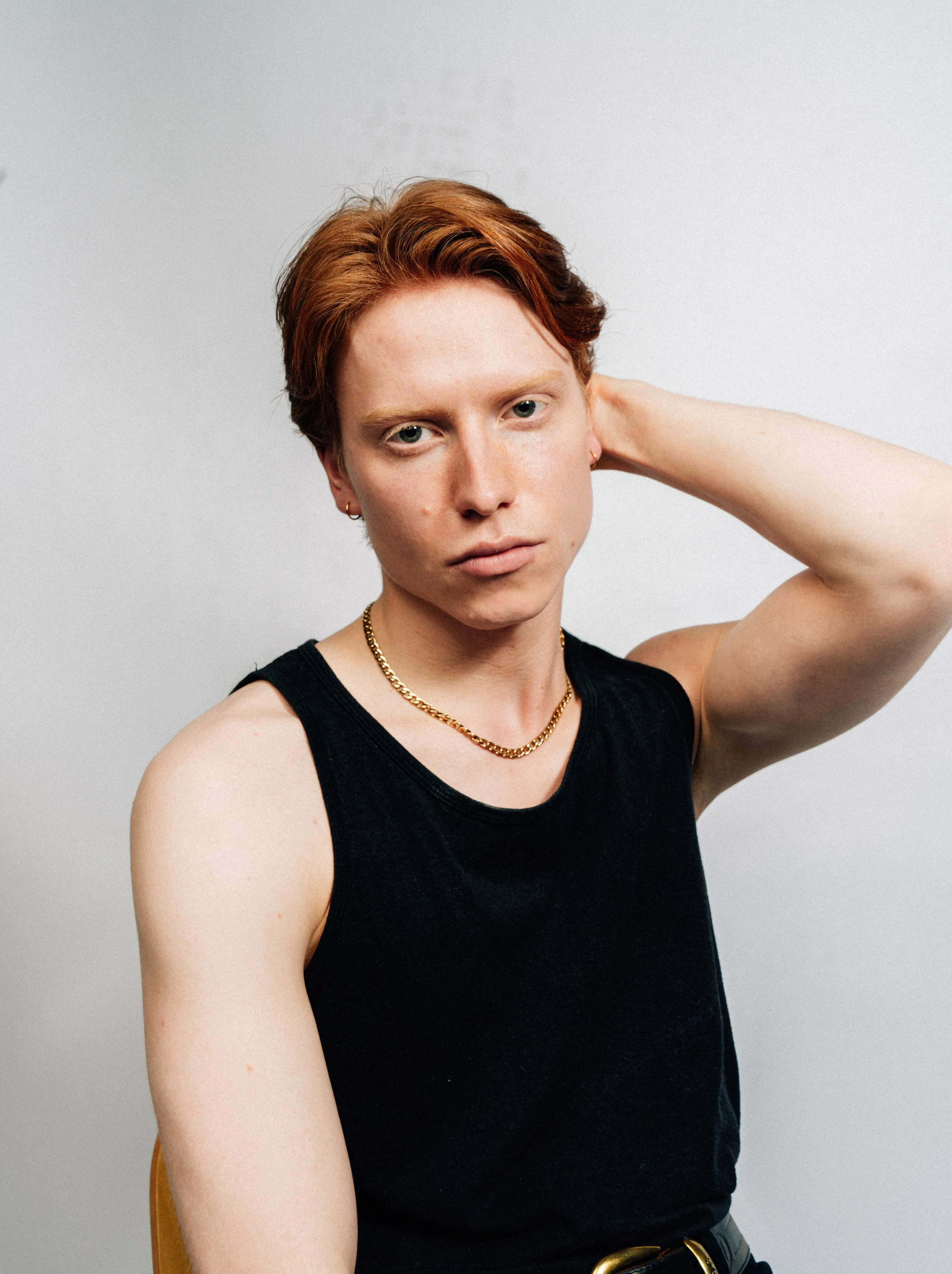 Porträt einer Person mit kurzen roten Haaren, schwarzem Tanktop, Goldkette, sitzt vor grauem Hintergrund, Blick in die Kamera, wird den Kopf mit einer Hand berührt.