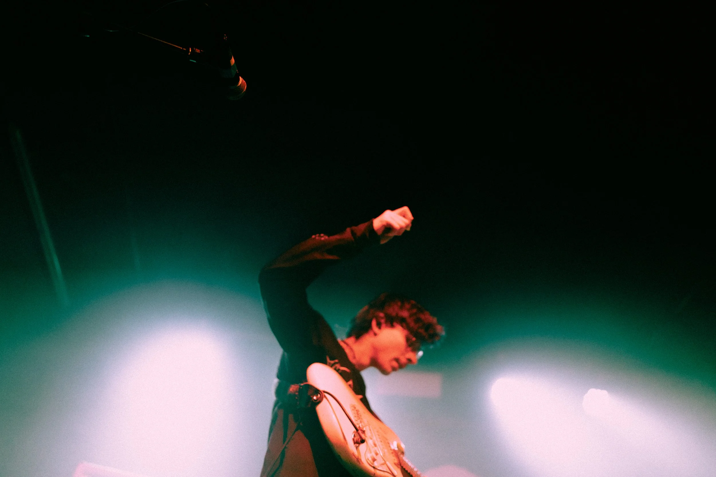 Musiker bei einem Auftritt, der eine E-Gitarre spielt und die Faust in die Luft streckt, auf einer Bühne mit bunten Lichteffekten.