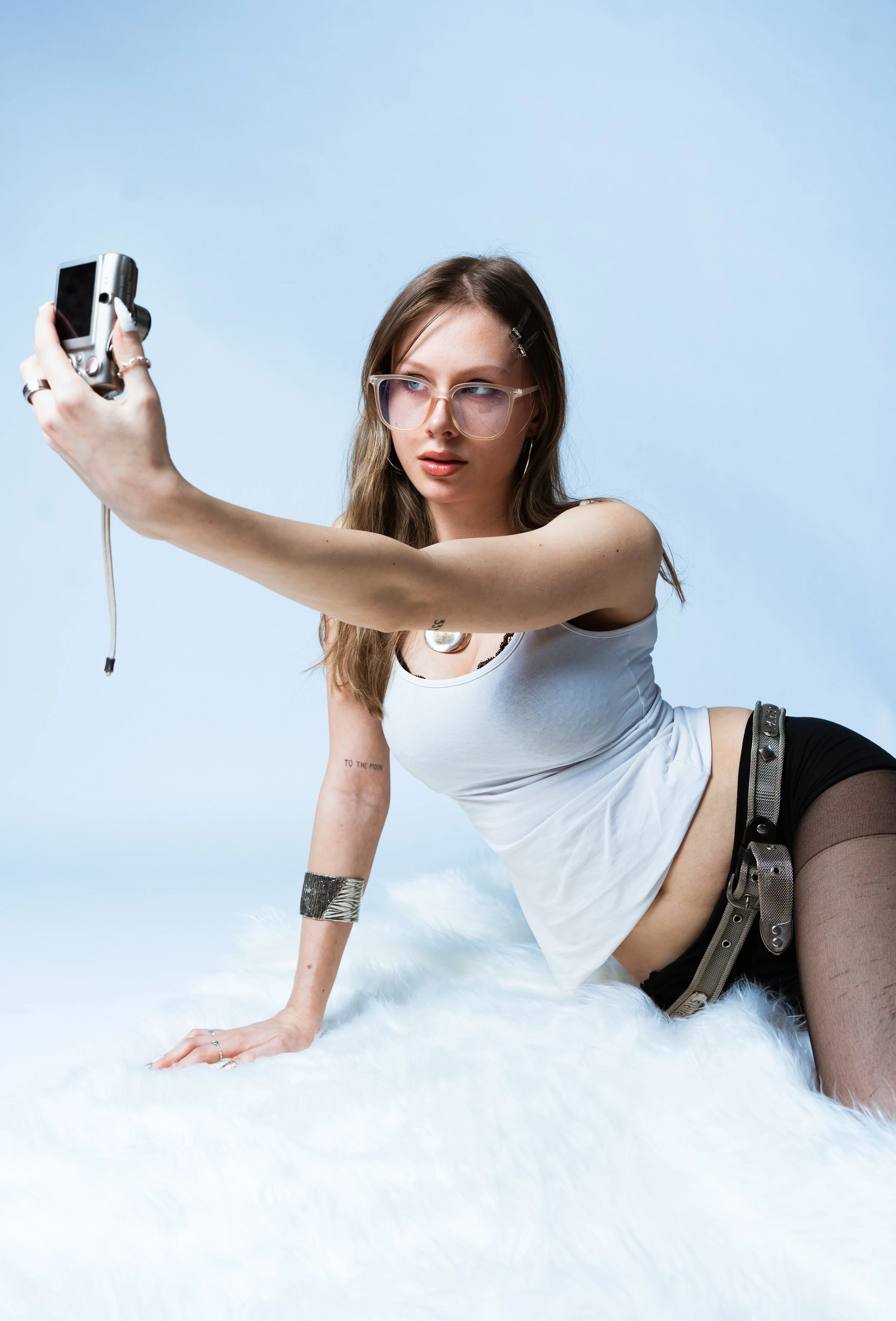Junge Frau macht ein Selfie mit einer Digitalkamera, liegt auf einer weißen Felldecke, trägt große Brille, weißes Top, schwarze Shorts, Strumpfhosen, und hat verschiedene Schmuckstücke.