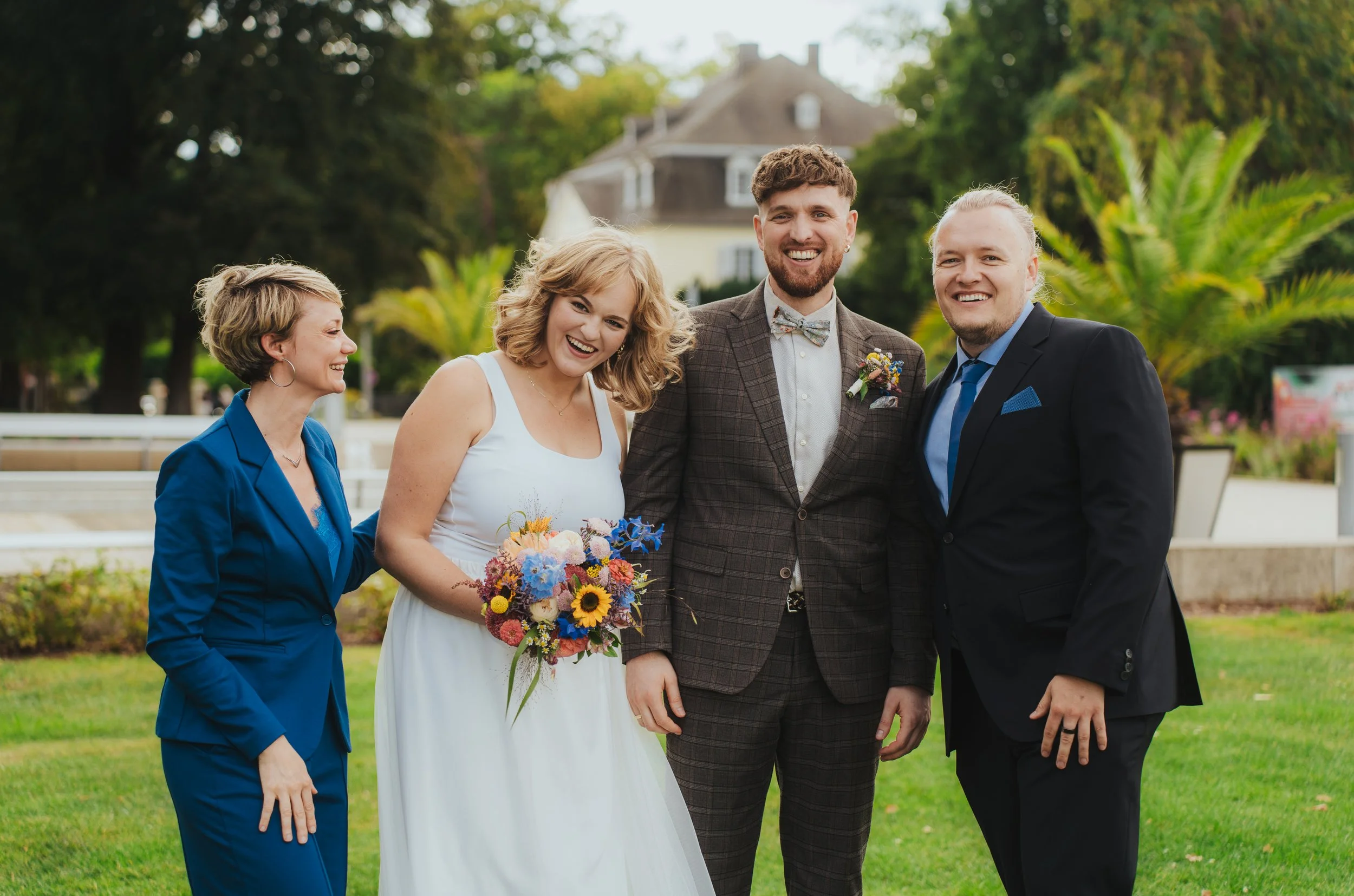Vier Personen, darunter eine Braut in Weiß mit Blumenstrauß und ein Bräutigam in Anzug, lachen und posieren im Garten bei einer Hochzeit.