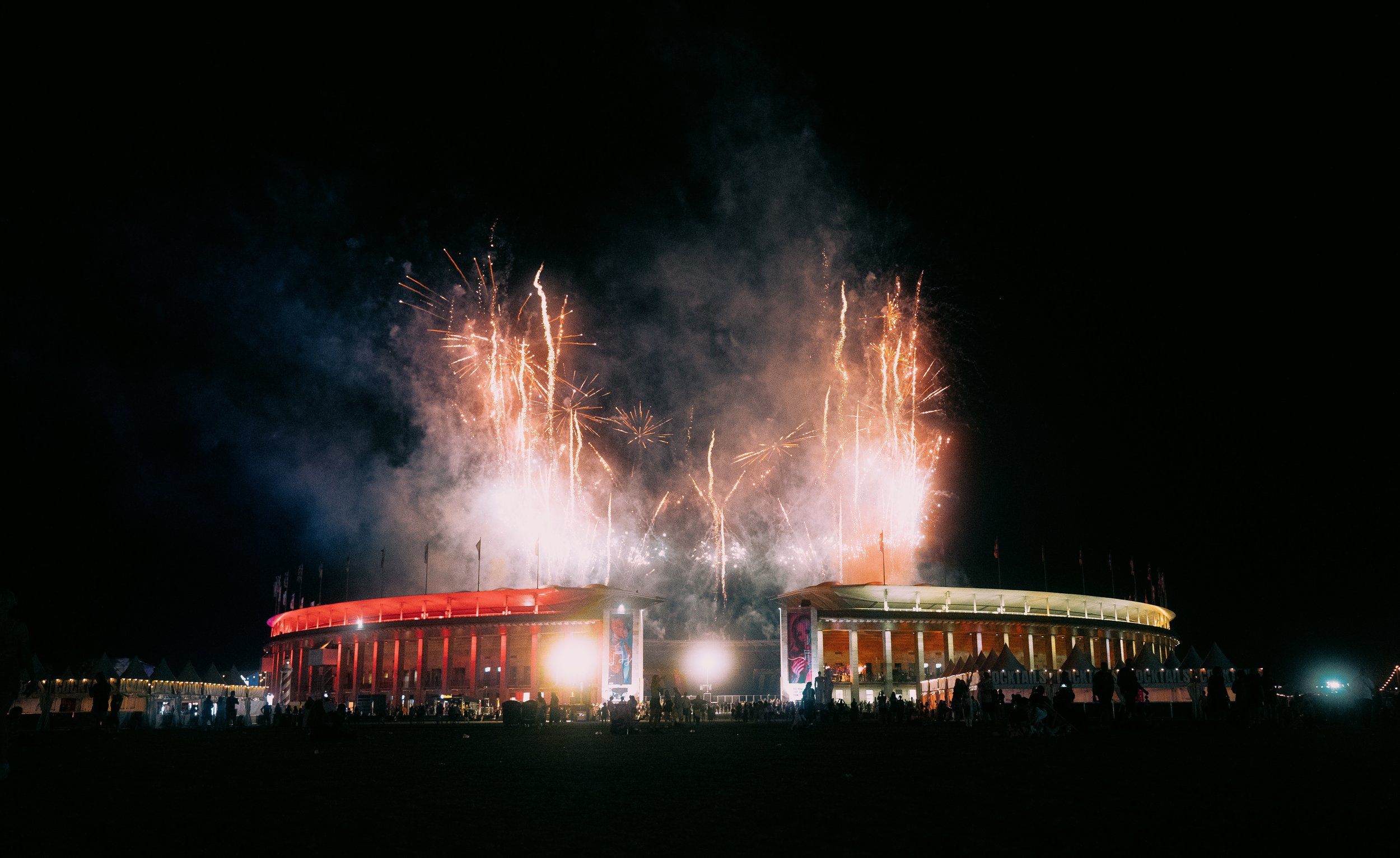 Feuerwerk über einer Veranstaltungsarena bei Nacht, mit Menschen im Vordergrund.