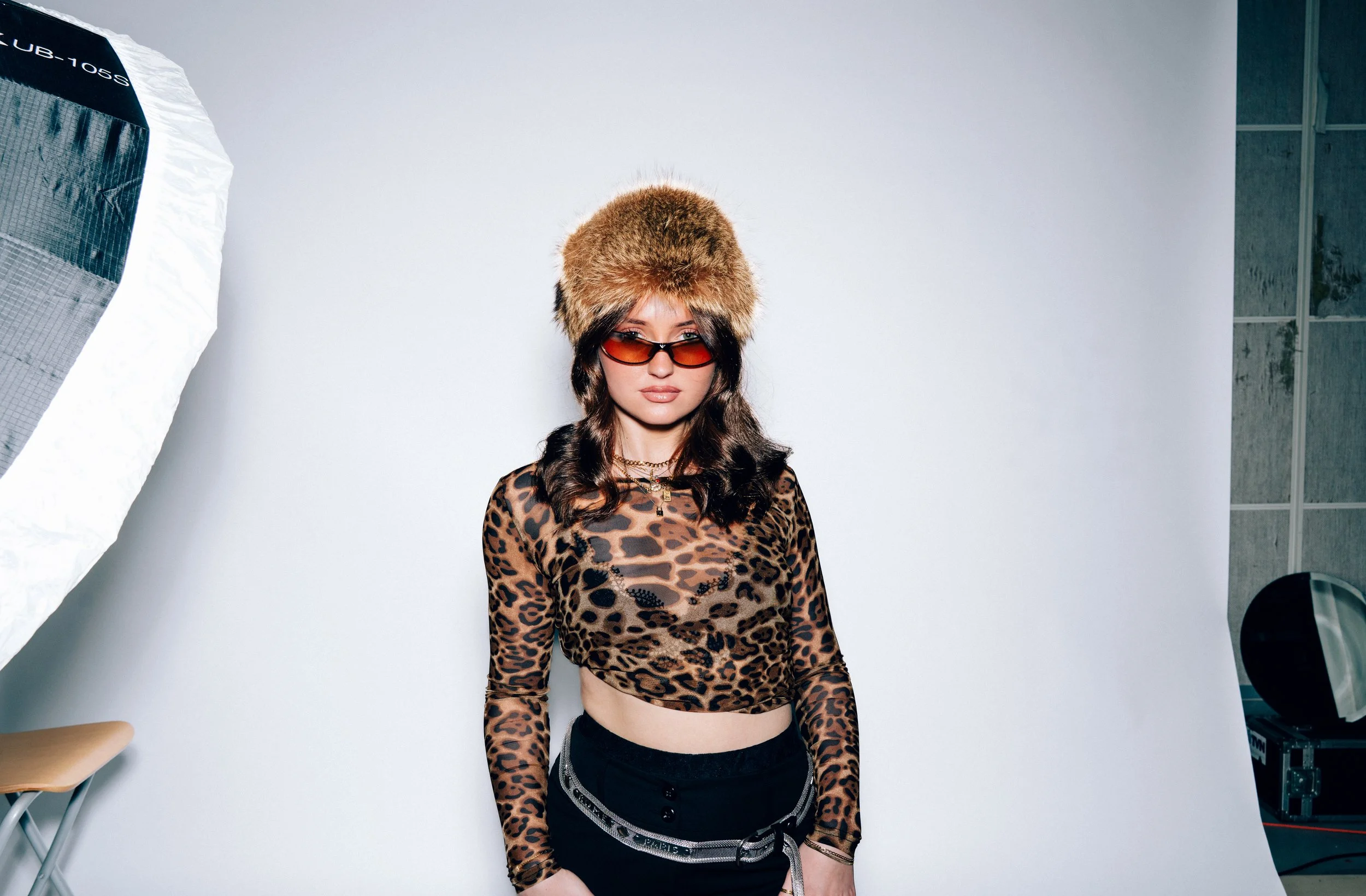 Junge Frau im Fotostudio mit Leo-Muster-Shirt, Kapuzenpullover, Sonnenbrille, und XXL-Fellmütze.