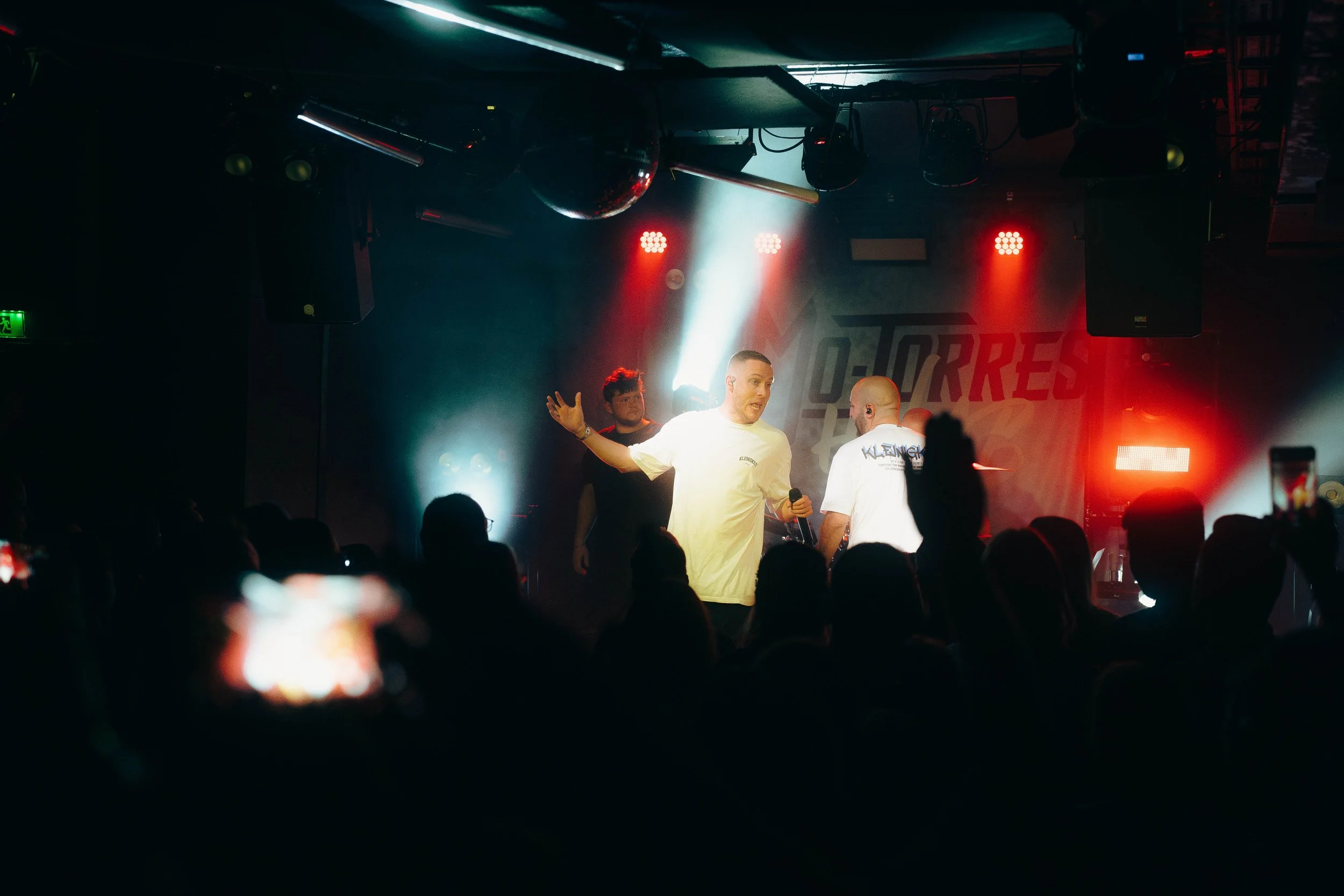 Eine Gruppe von Menschen auf einer Bühne bei einer Live-Performance, mit einem Sprecher, der mit einem Mikrofon spricht, während die Zuschauer im Vordergrund sitzen und das Geschehen beobachten.