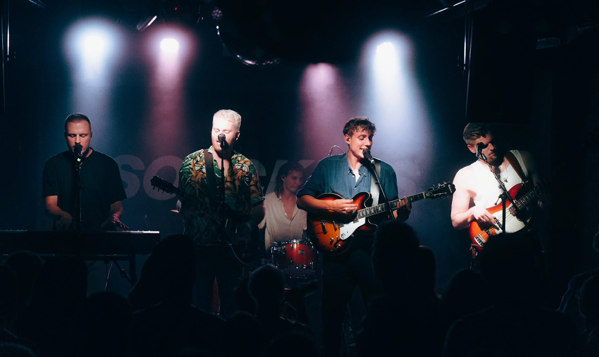 Eine Band mit vier Mitgliedern spielt auf einer Bühne bei einem Live-Konzert, mit Lichtstrahlen und Publikum im Vordergrund.