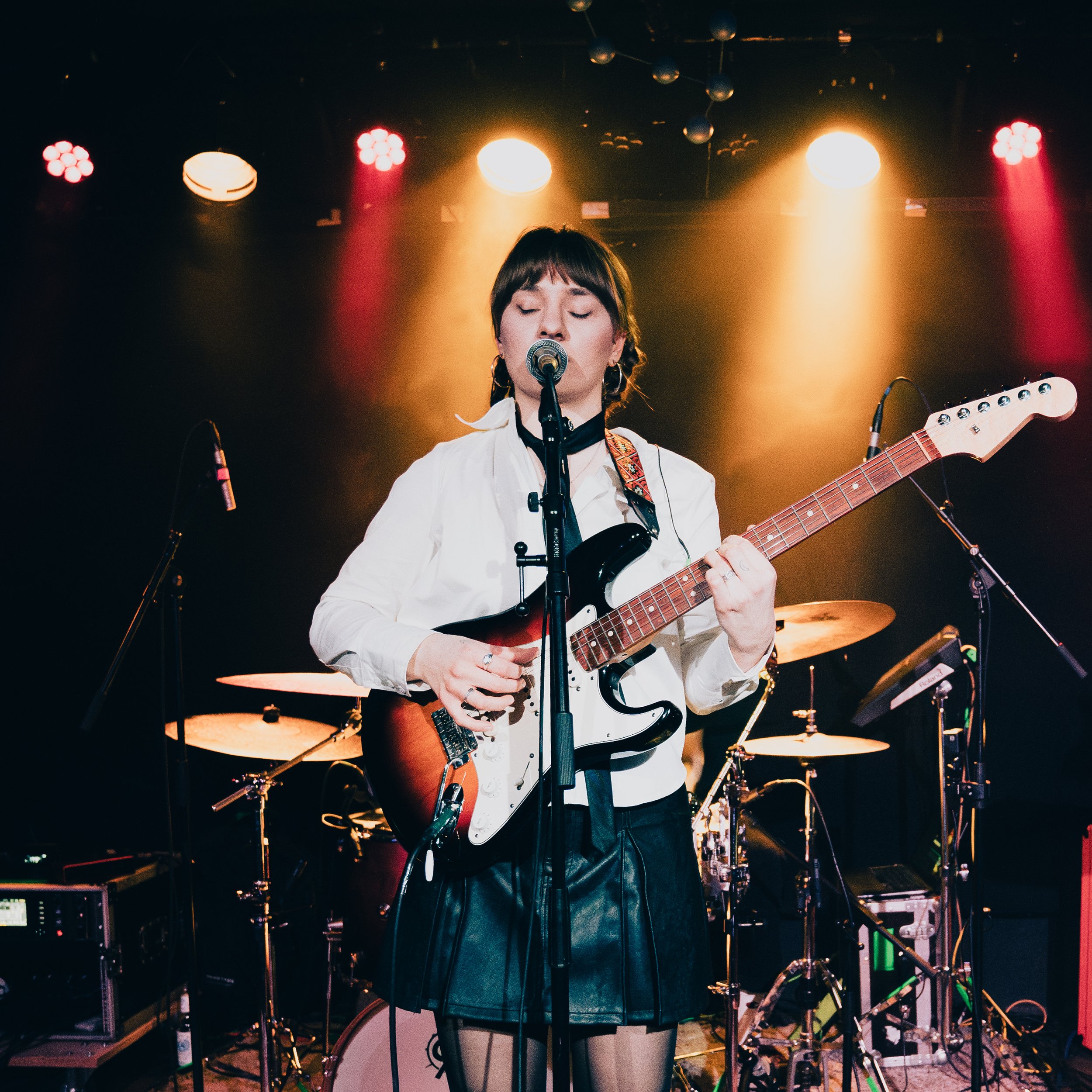 Junge Frau spielt Gitarre und singt bei einem Live-Konzert auf der Bühne, im Hintergrund sind Schlagzeug und andere Musikinstrumente sichtbar, mit bunten Bühnenlichtern.