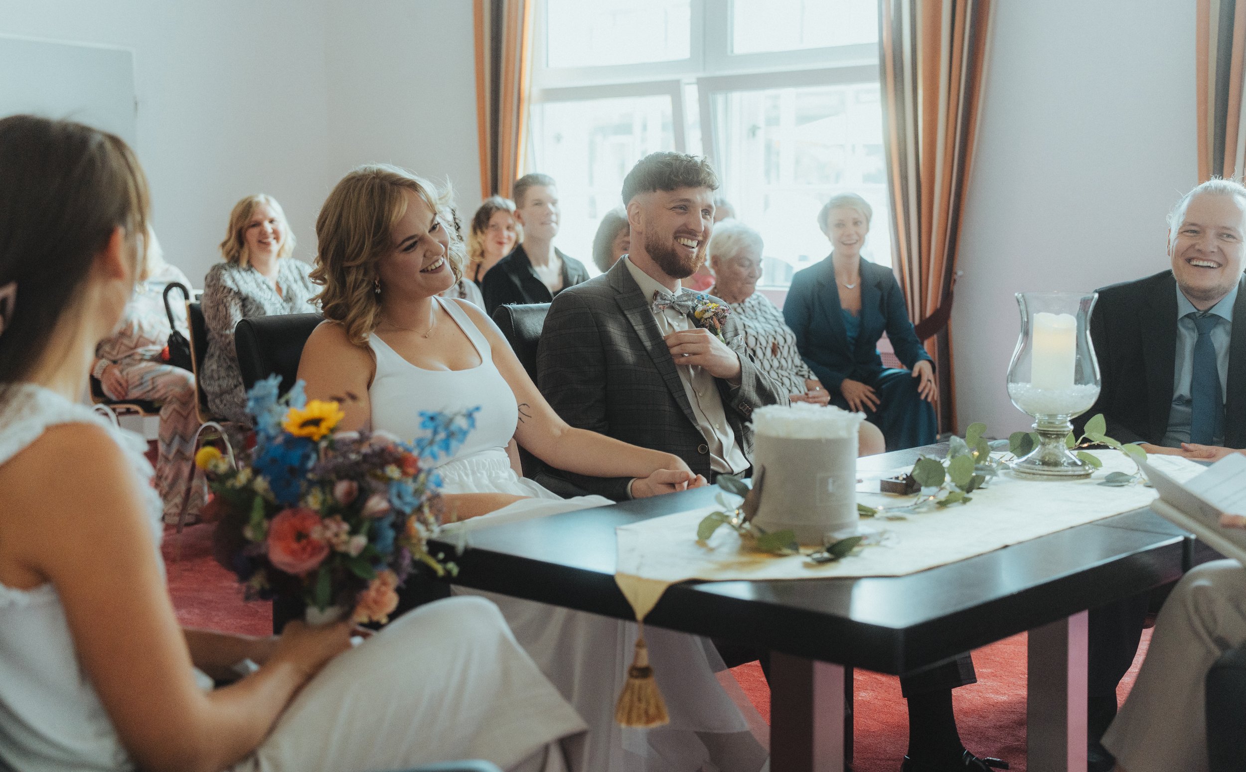 Menschen bei einer Hochzeitsfeier sitzen um einen Tisch, lachen und haben Freude, mit Blumen und Kerzen dekoriert.