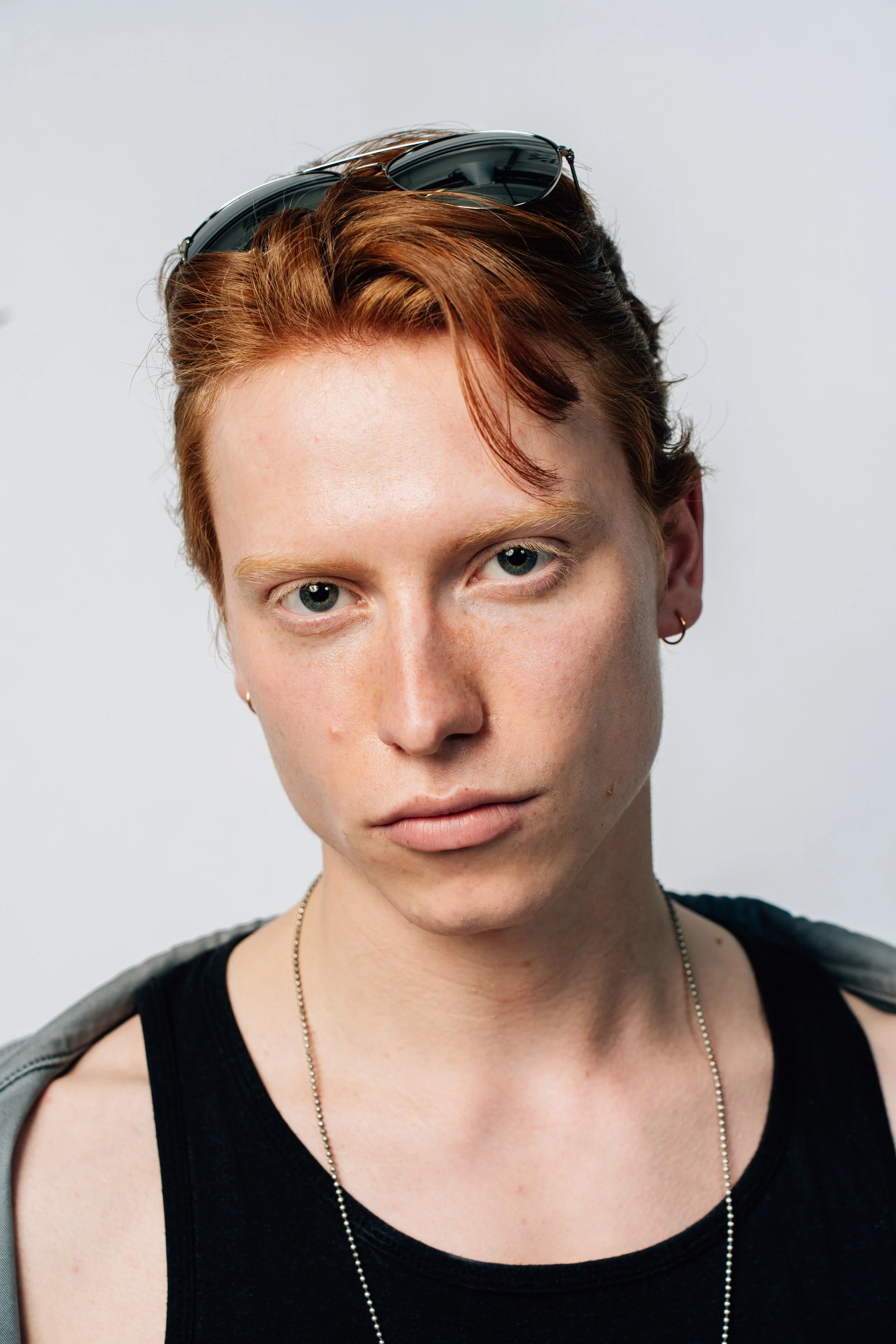 Nahaufnahme eines jungen Mannes mit roten Haaren, Sonnenbrille auf dem Kopf, schwarzen Tanktop und silberner Halskette vor einem weißen Hintergrund.
