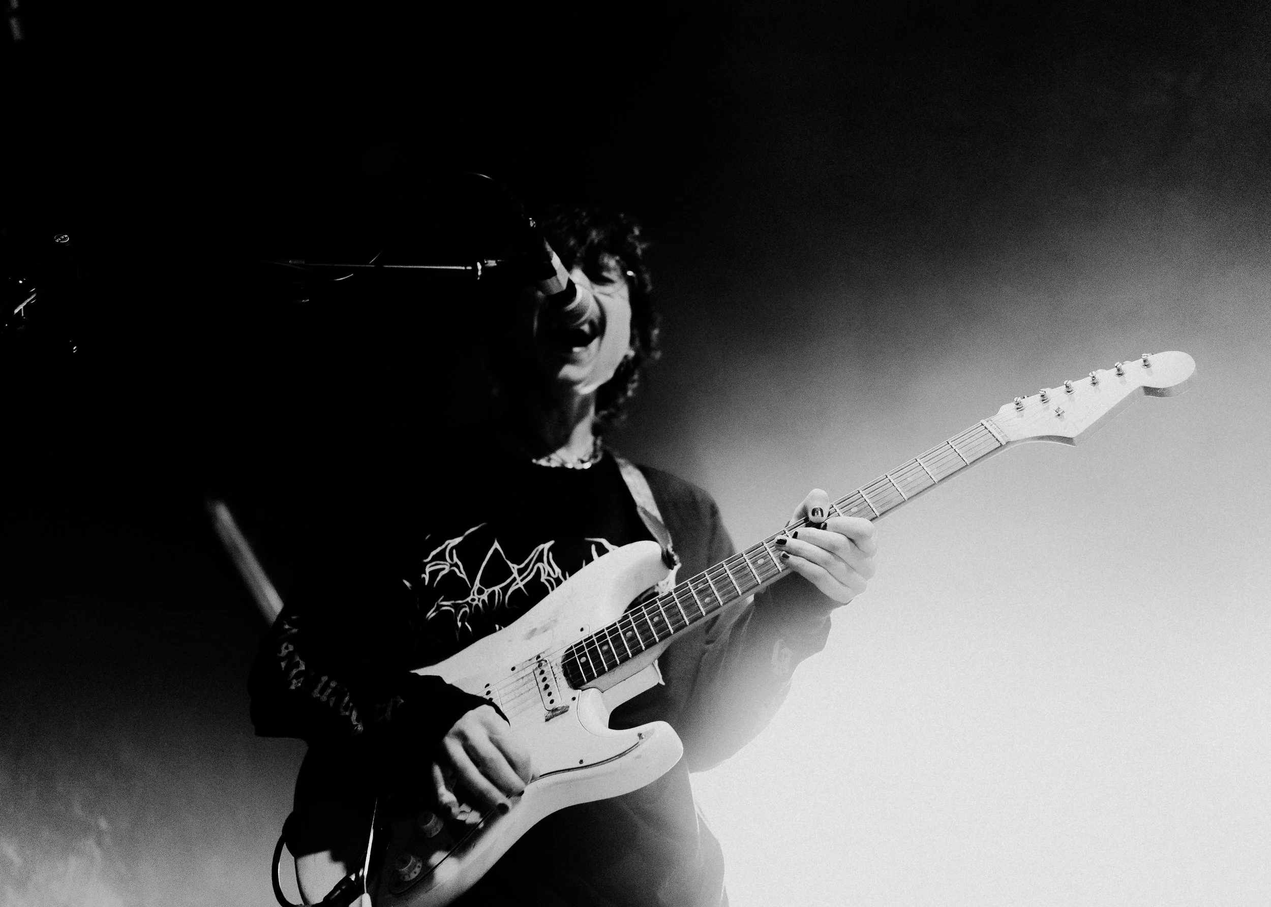 Ein Musiker spielt eine E-Gitarre auf einer Bühne, er singt in ein Mikrofon, trägt ein dunkles T-Shirt mit einer Band-Logo, die Bühne ist schwach beleuchtet, das Bild ist schwarz-weiß.