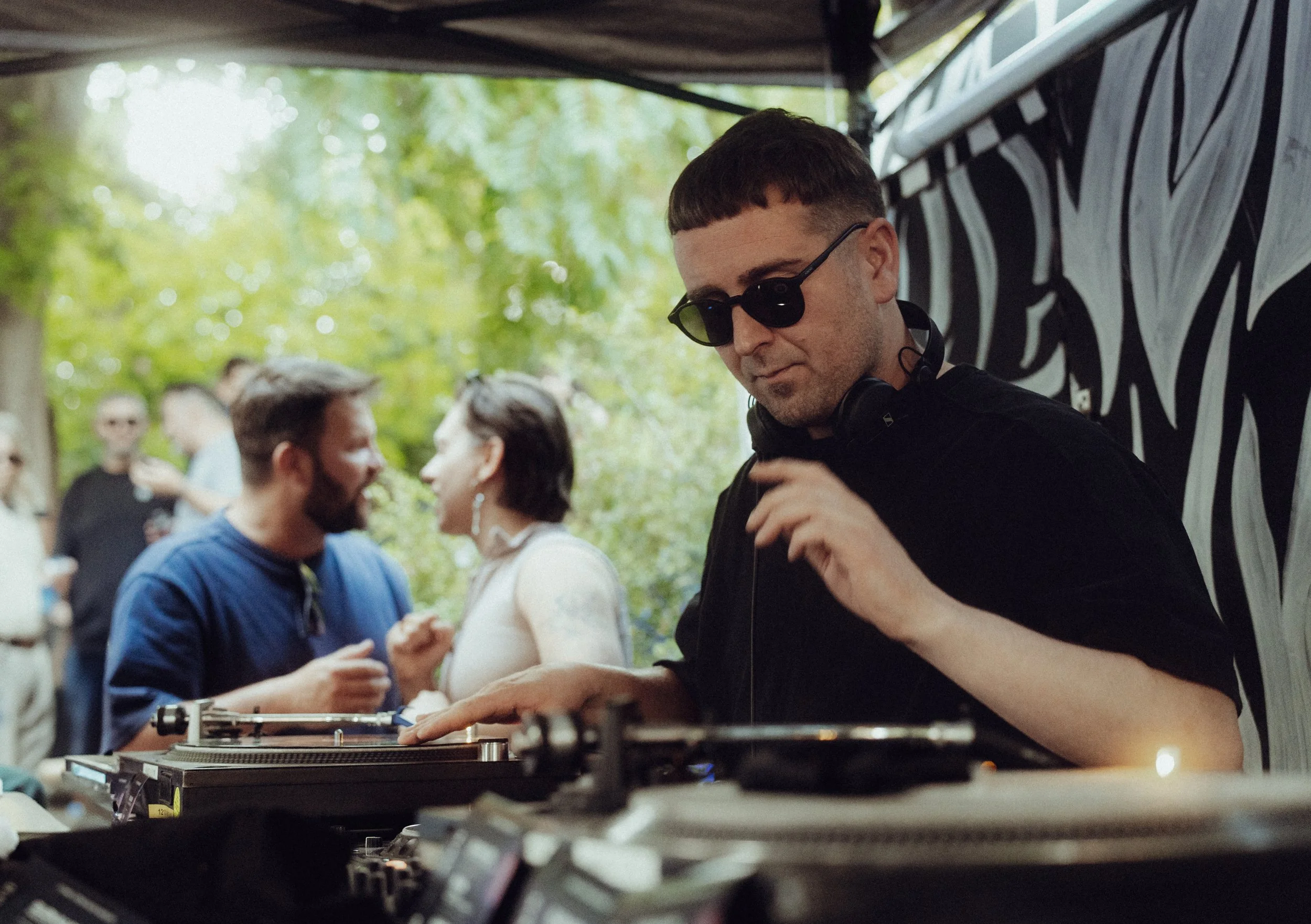 Ein DJ mit dunklen Sonnenbrillen und Kopfhörern hinter einem Mischpult, im Hintergrund feiern Menschen bei einem Outdoor-Event in der Natur.