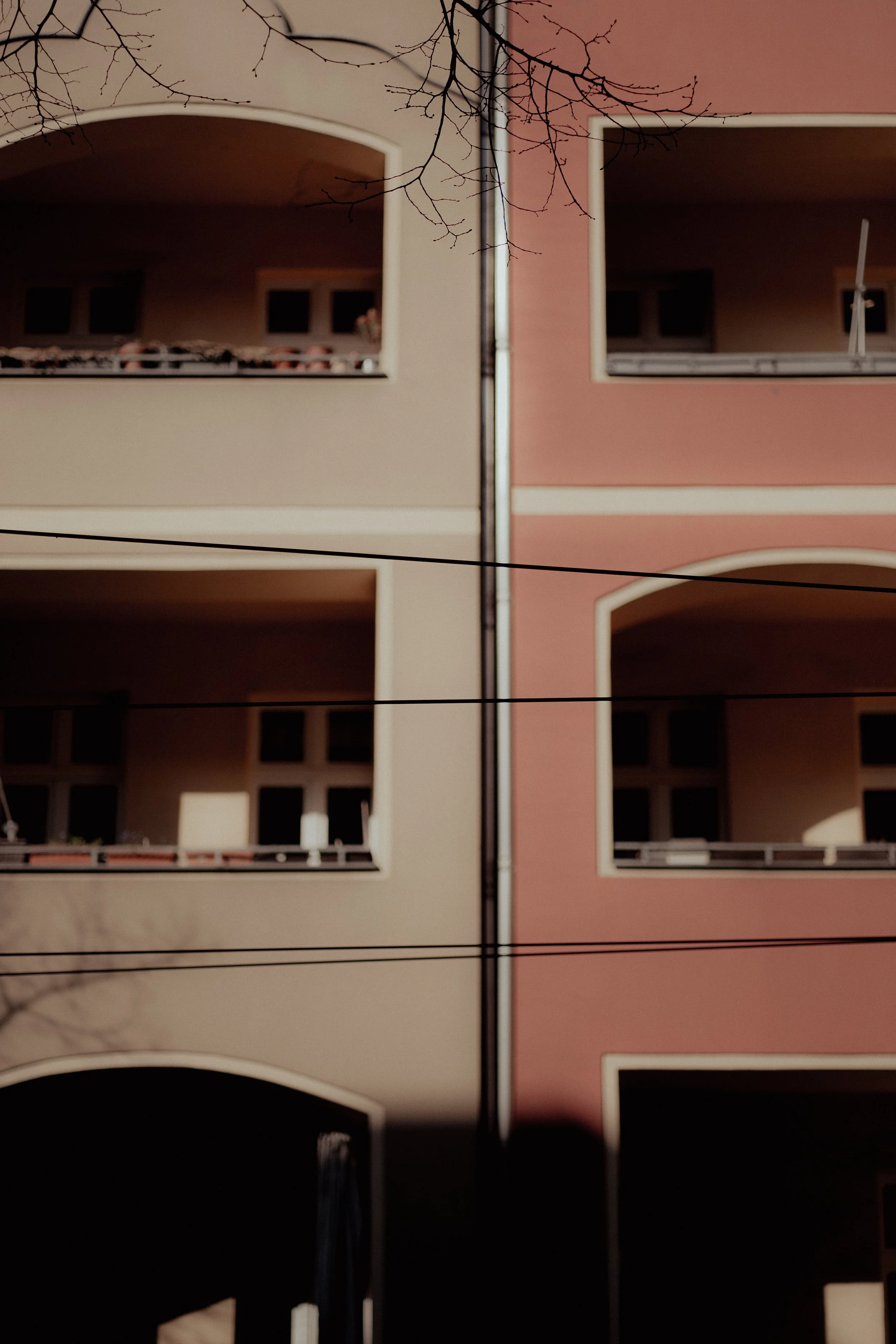 Ansicht zweier nebeneinanderstehender Mehrfamilienhäuser, eines in beige, eines in rosa, bei Nacht, mit sichtbaren Balkonen und Fenstern, einige Kabel verlaufen vor den Häusern, kahle Baumzweige im oberen Bereich.