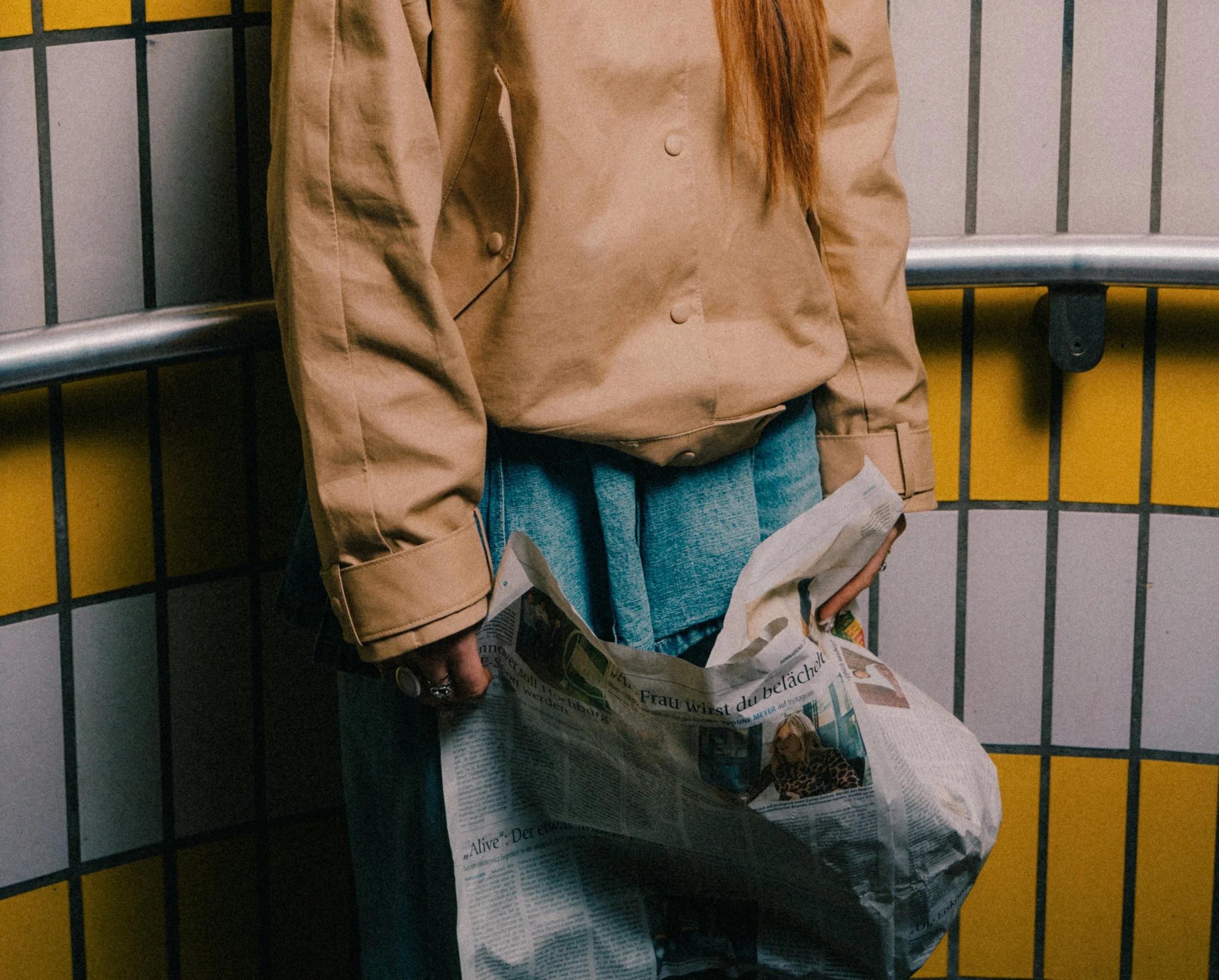 Person steht in einer Bahn- oder U-Bahn-Station mit gelb-schwarz gestreiften Wänden, hält eine Zeitung in der Hand.