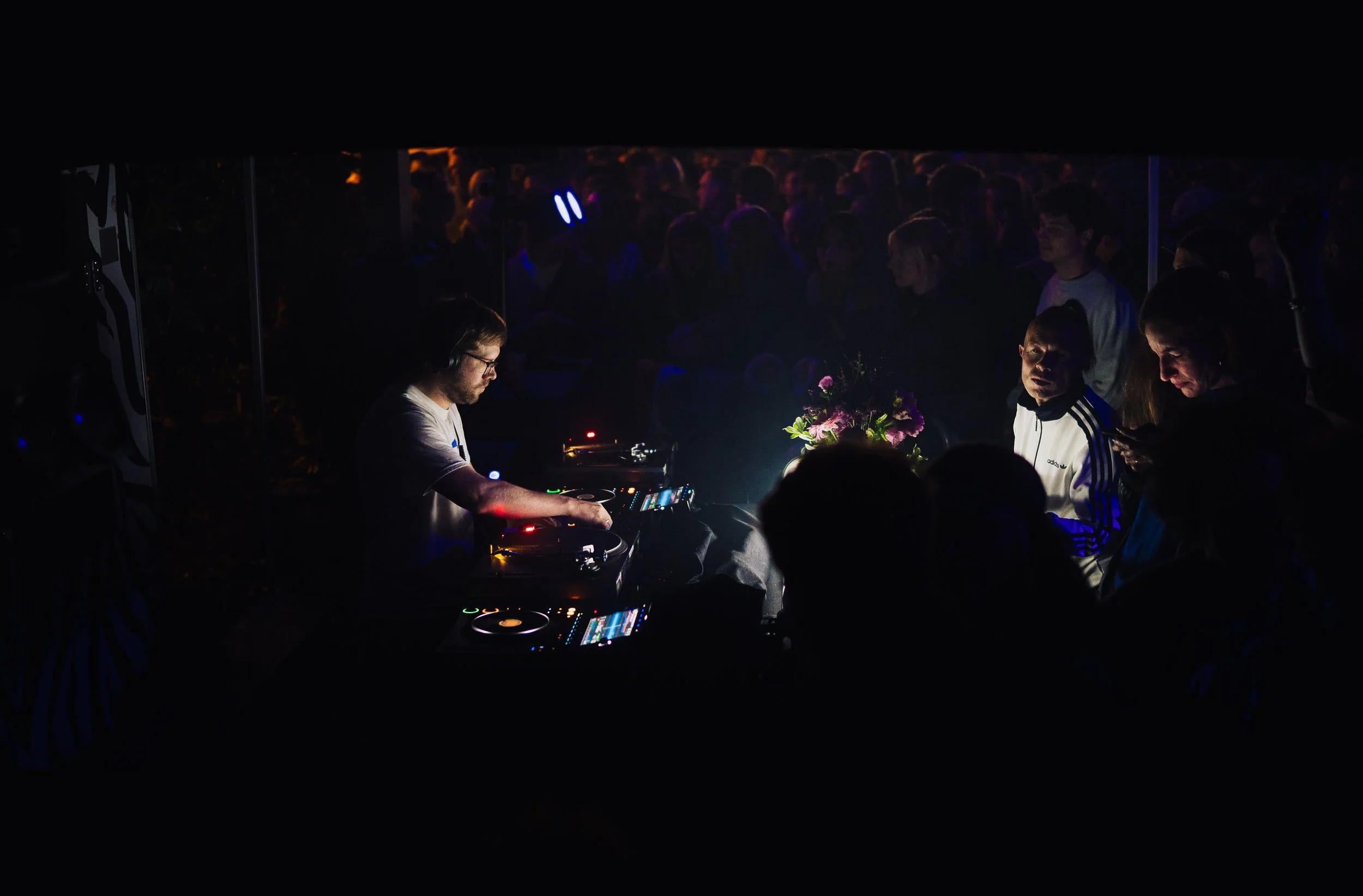 DJ mit Kopfhörer spielt auf einem DJ-Deck in einer dunklen, mit Publikum gefüllten Umgebung, beleuchtet von blauem und rot-rosa Licht, während einige Zuschauer auf der rechten Seite das Smartphone benutzen.