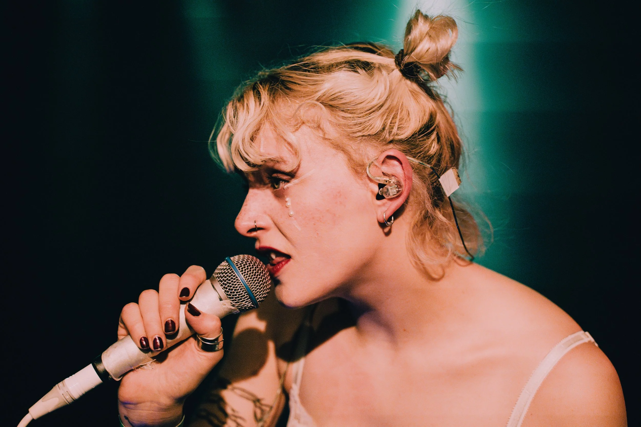 Junge Frau singt in einem Mikrofon, trägt mehrere Ohrringe und ein randloses Ohrrad, hat blaue Frisur, Locken und trägt ein Tanktop, mit Tattoos am Arm, im dunklen Licht bei einem Auftritt.