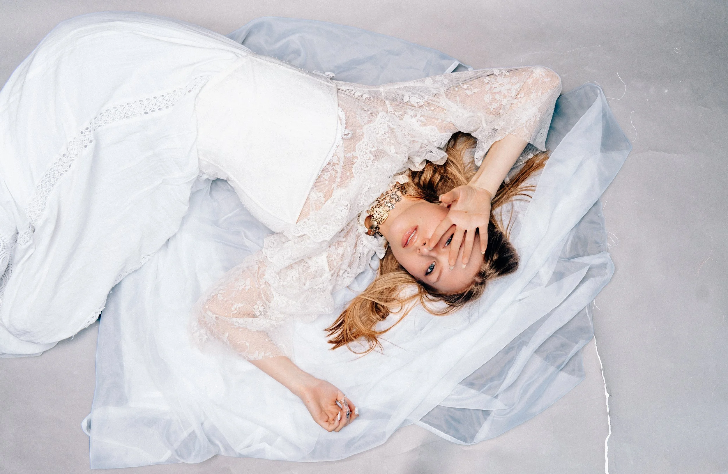 Frau liegt auf weißem Bett, trägt ein transparentes Kleid mit Spitzenmuster und eine große Halskette, hält eine Hand vor das Gesicht, blickt in die Kamera.