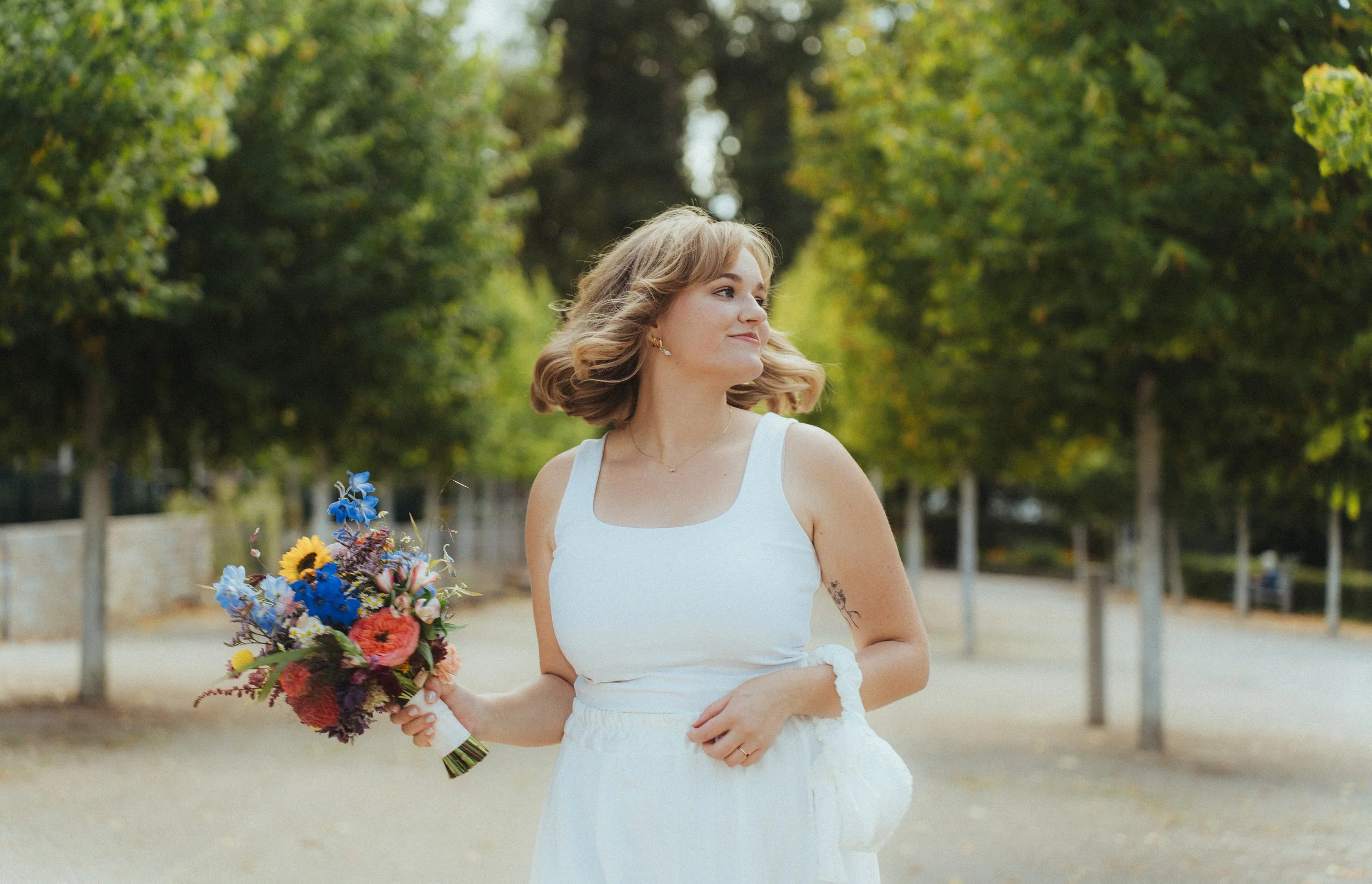 Eine junge Frau im weißen Kleid hält einen Blumenstrauß und steht in einer Parkumgebung mit Bäumen.