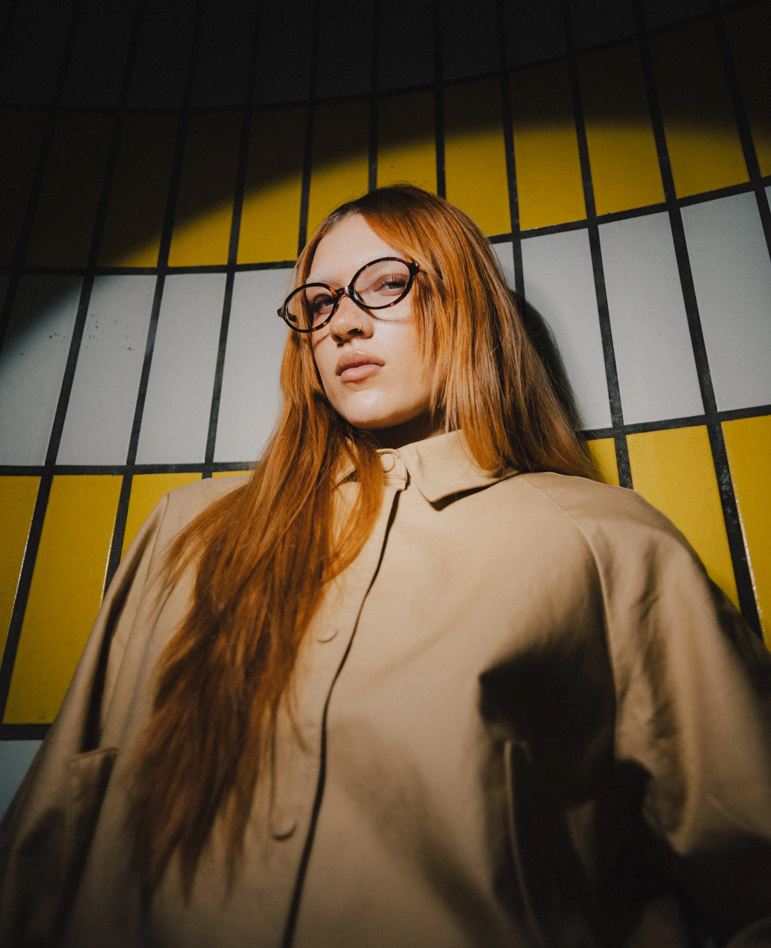 Junge Frau mit roten Haaren, Brille und Nasenpiercing steht vor einer bunten Wand mit schwarz-weißen und gelben Quadraten, teilweise im Schatten.