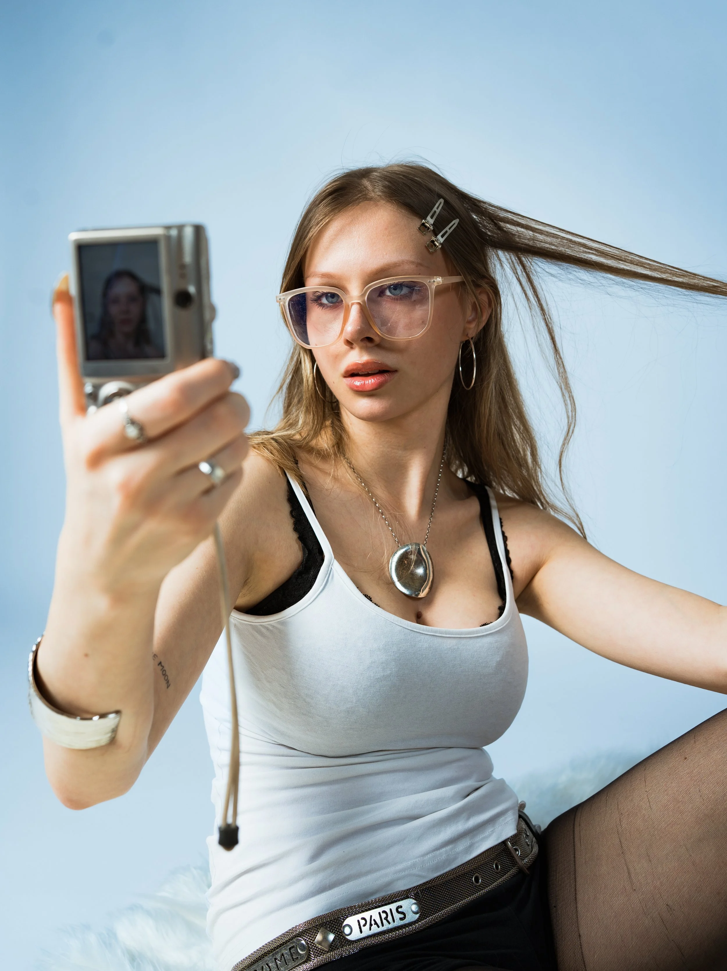 Junge Frau mit Brille macht Selfie mit Digitalkamera, blauer Hintergrund, trägt weiße Top, schwarze Netzstrumpfhose, Schmuck und Haarclips.