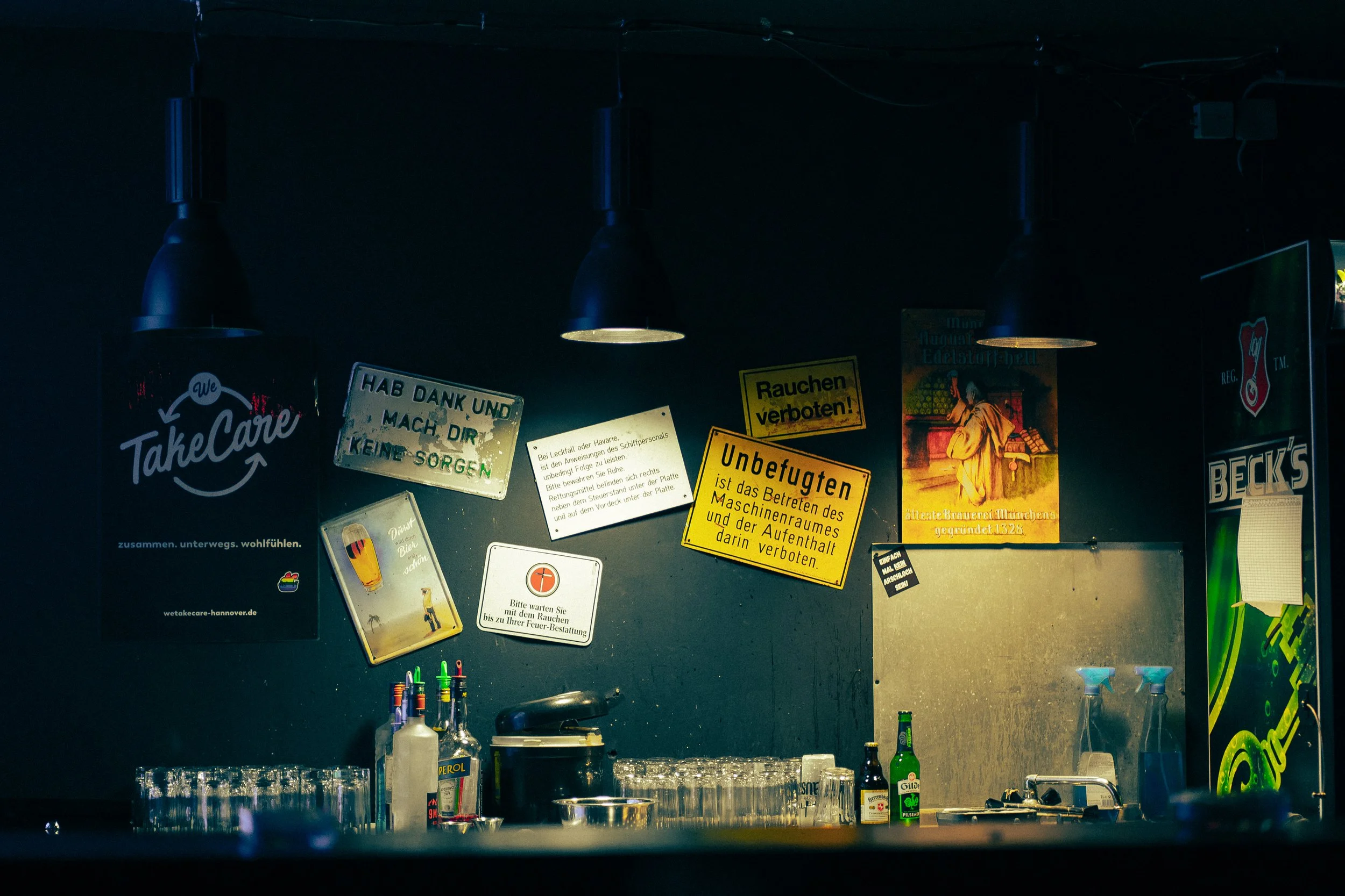 Blick auf eine Bar mit verschiedenen Postern und Hinweisschildern an der Wand, darunter ein Poster für TakeCare, diverse alkoholische Getränke, leere Gläser, Flaschen und Getränke, sowie einen Kühlschrank an der rechten Seite.