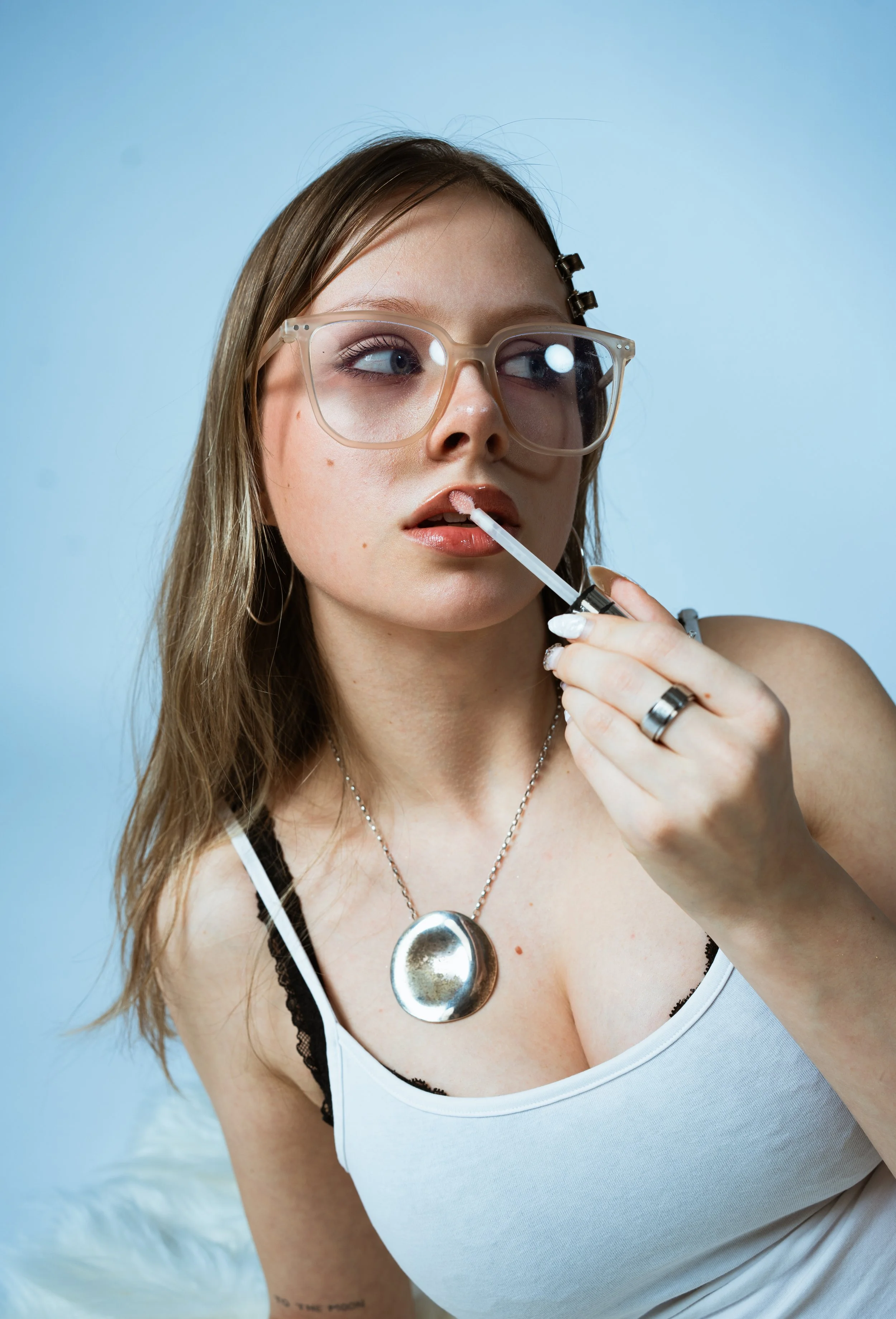 Junge Frau mit großen Brillengläsern trägt eine Halskette und einen weißen Top, während sie Lippenbinsel aufträgt.